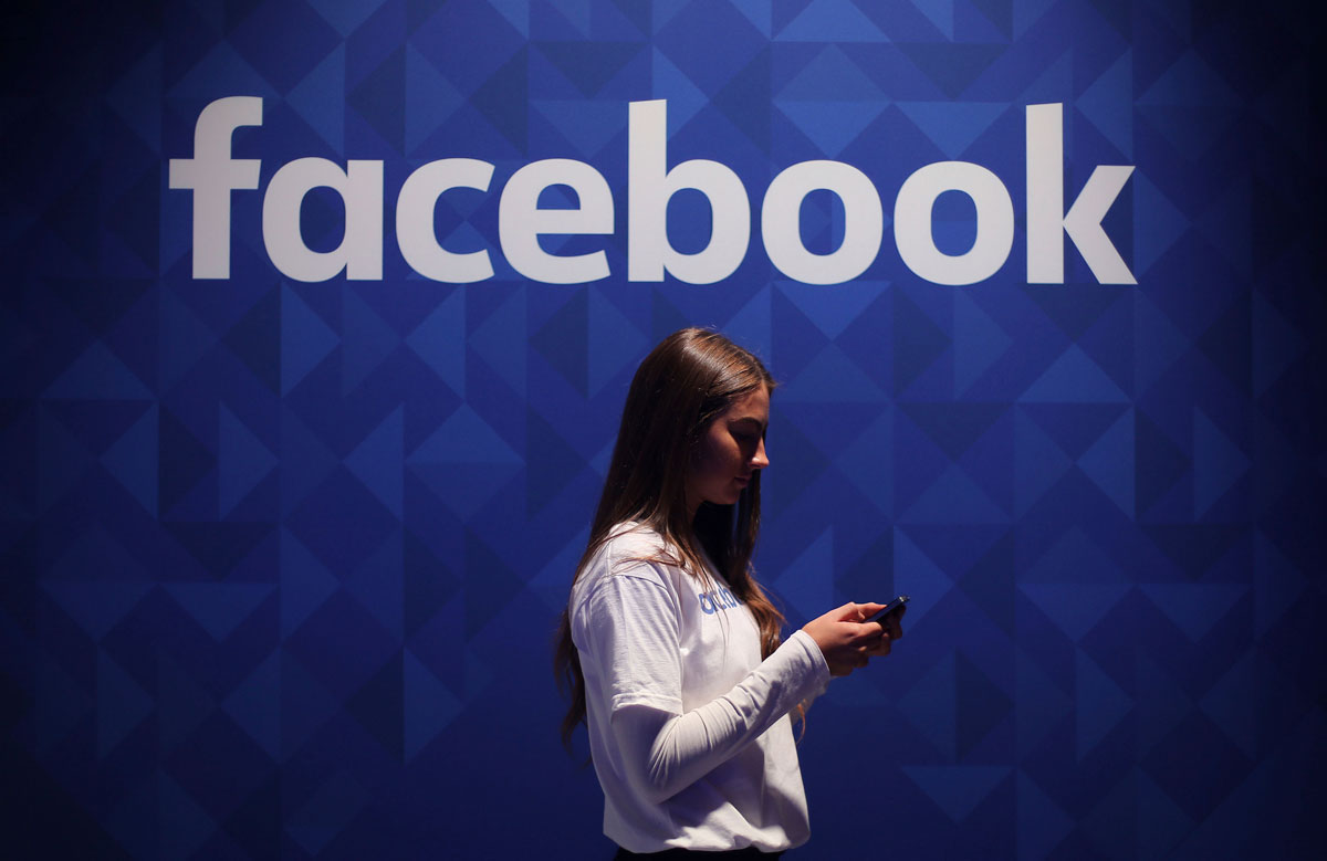 A woman looking at her smartphone beneath Facebook&#039;s logo