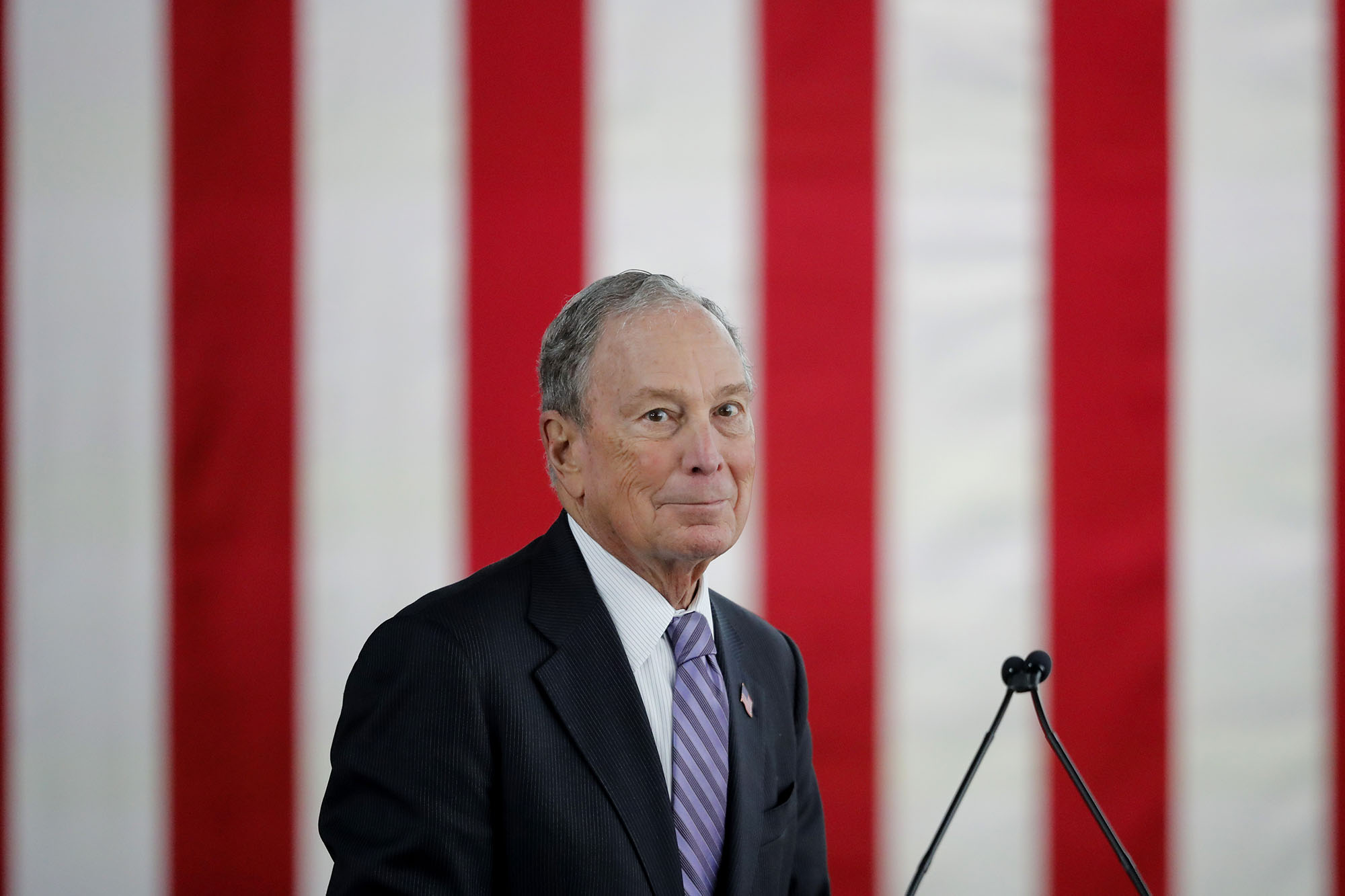 Michael Bloomberg, during a speaking event, with the stripes of the American flag in the background.