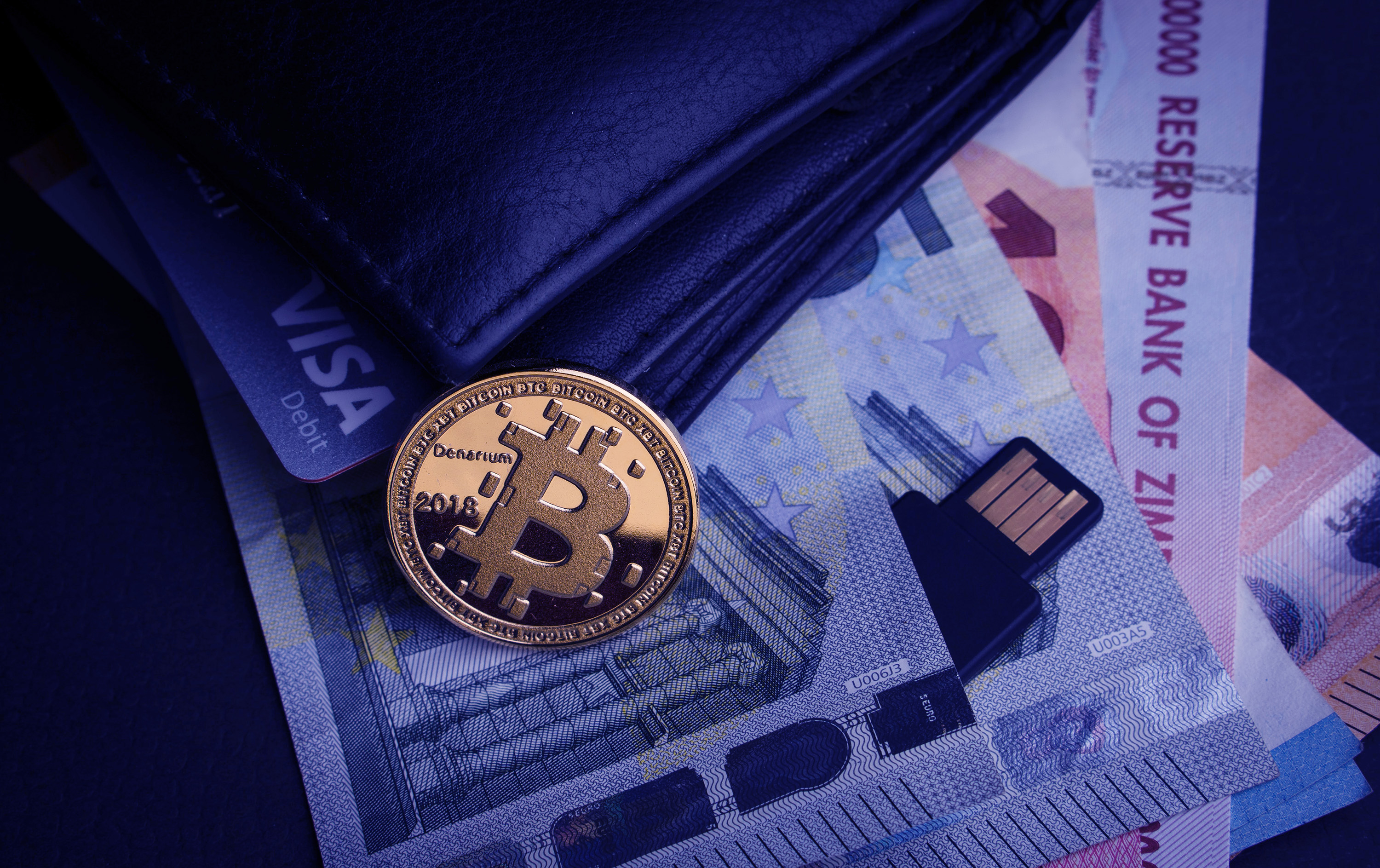 A physical representation of a bitcoin rests on a wallet that also contains a Visa card and paper banknotes from several countries.