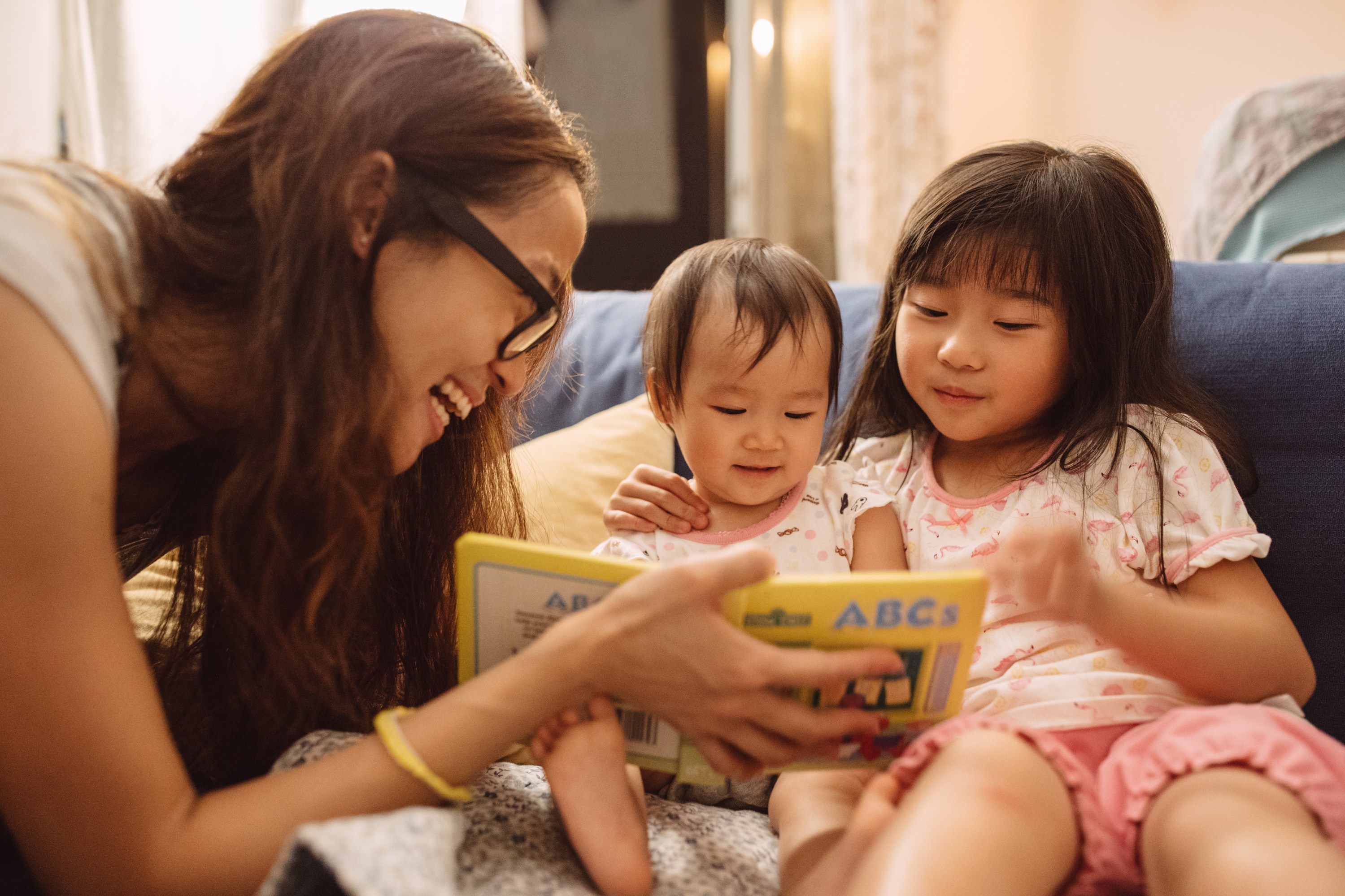 Mom reading story book to her daughter and baby on the couch.