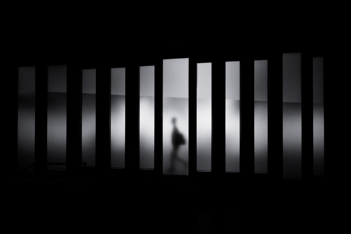 A person in shadows walks behind a series of windows in a black-and-white image