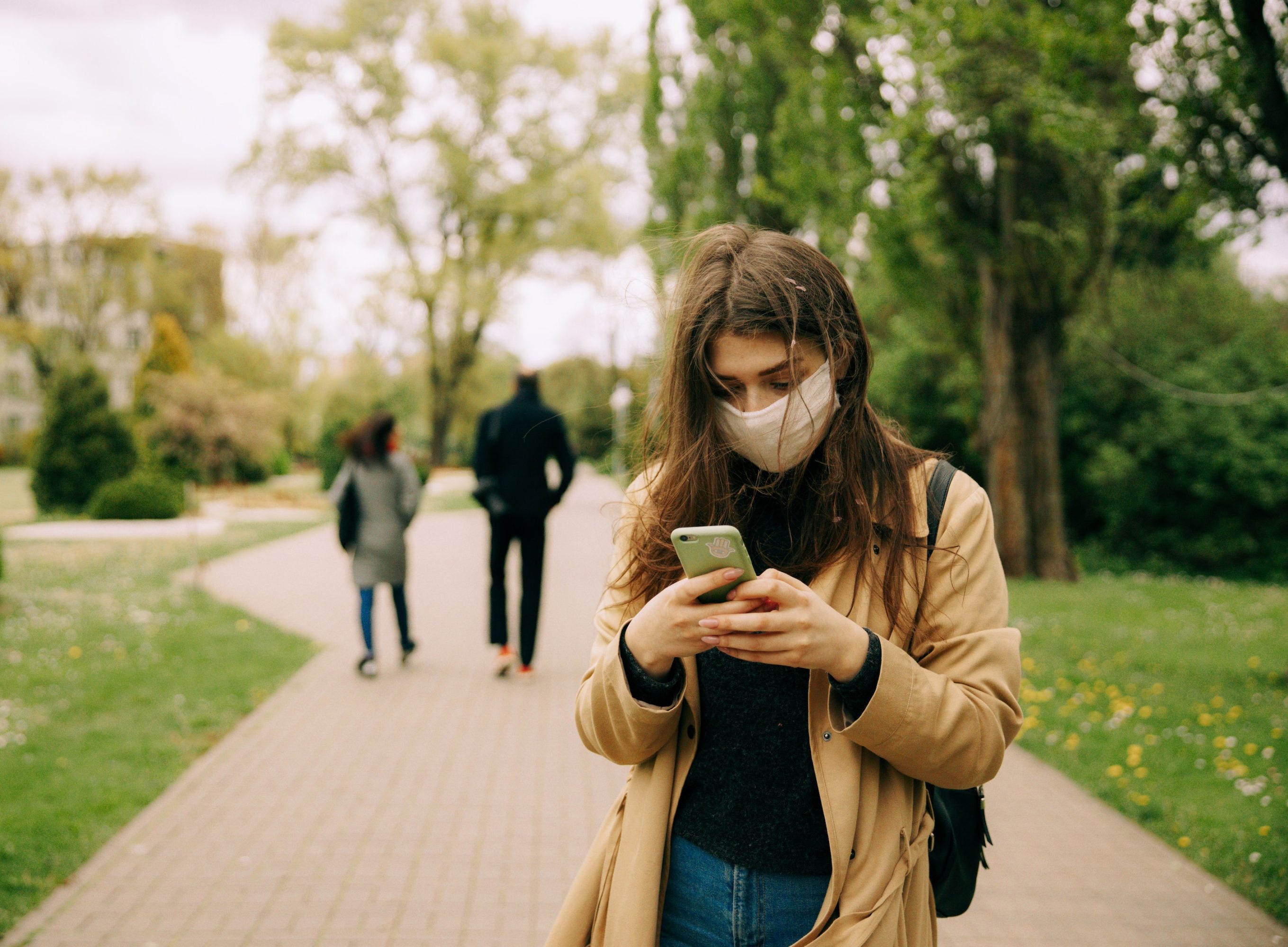 Woman on smartphone wearing face mask, with two people walking away in the background.