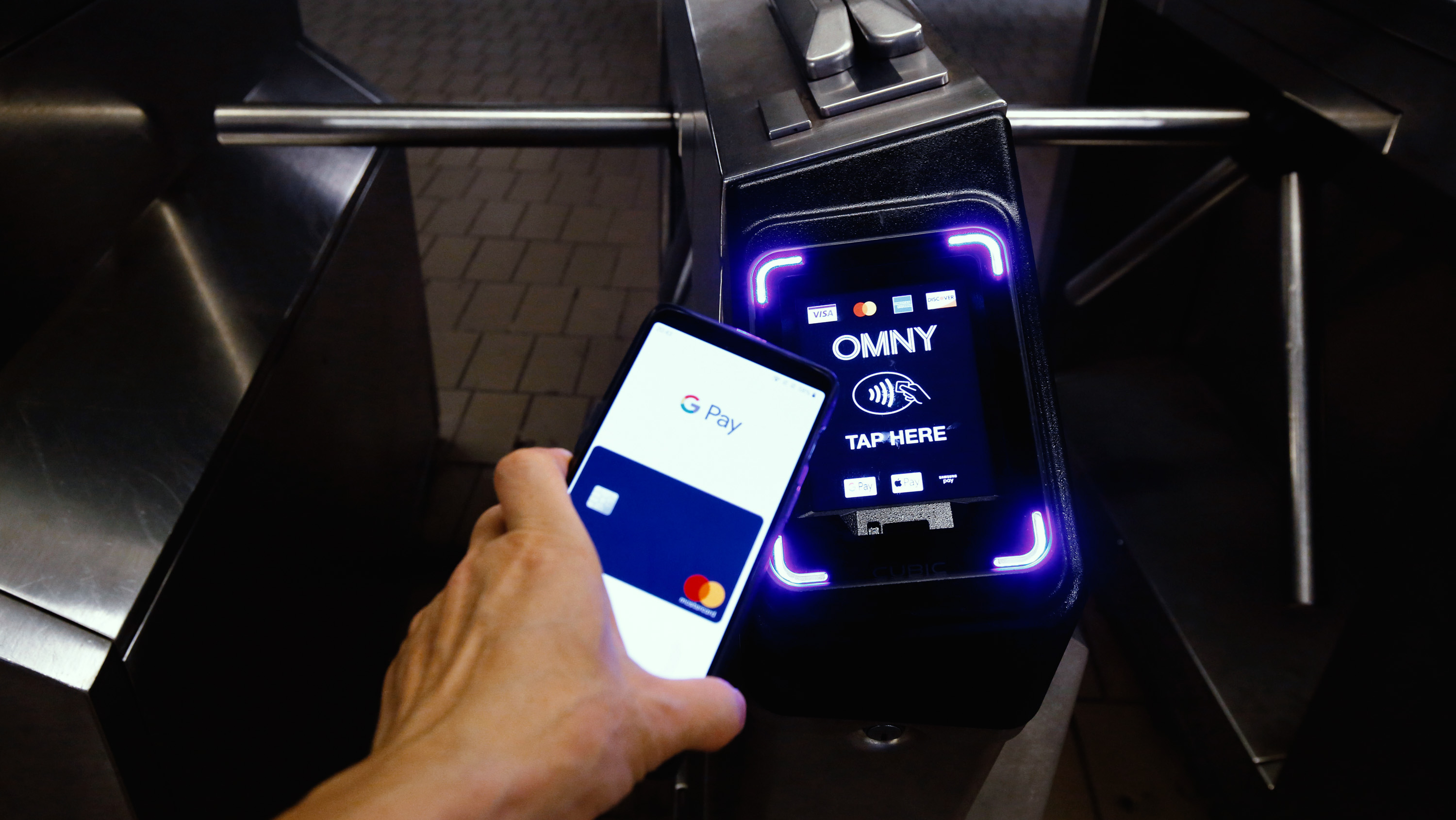 A smartphone with Google Pay taps a NYC Subway turnstile retrofitted with a contactless payment reader