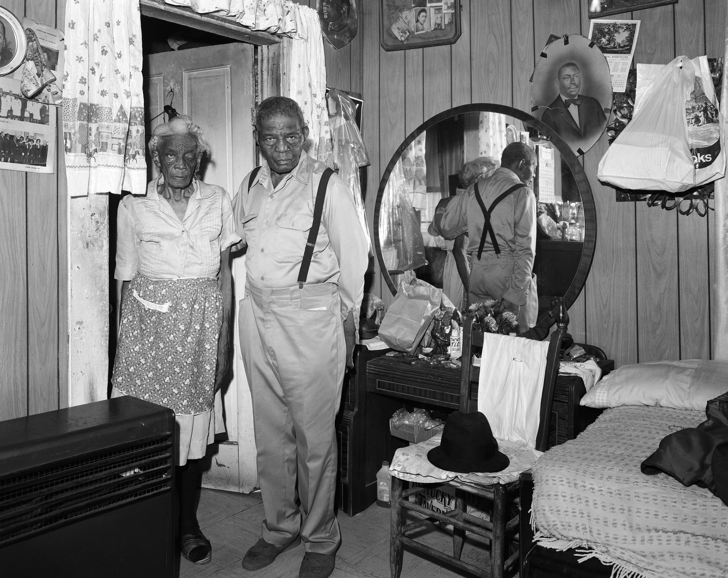 A old man and old woman, who is missing an eye, stand in the doorway of a room with a mirrored dresser, family photos, and a chair.