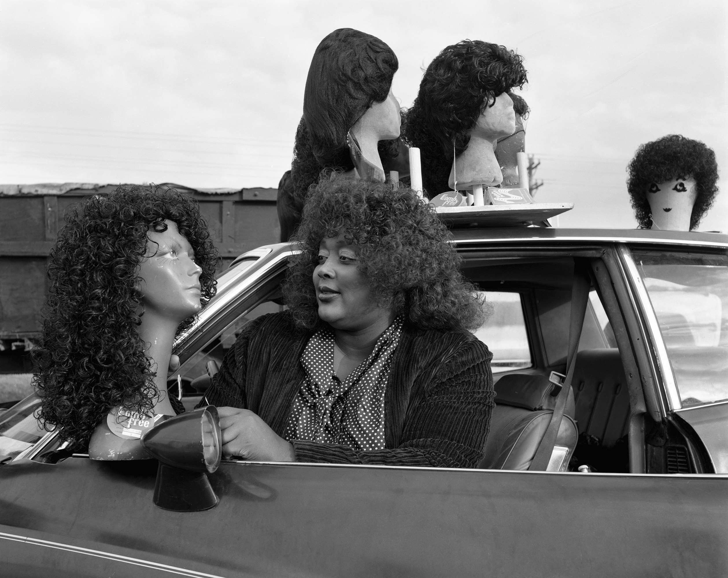 A woman leans out of the window of her car. Several mannequin heads with wigs are mounted to the roof, and she is holding one near her driver's side mirror.