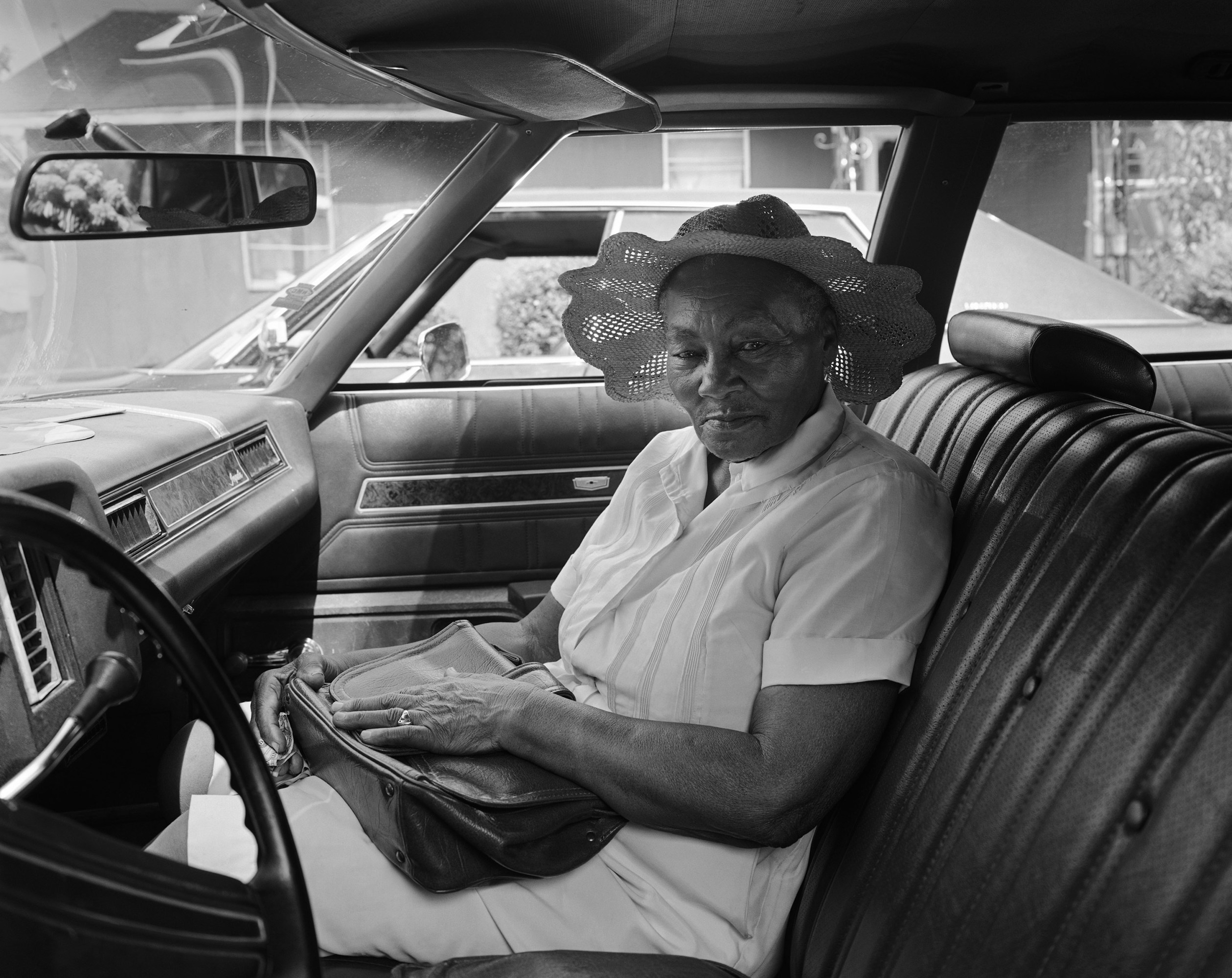 Driver's side view of the interior of a car where an older woman wearing a sunhat sits in the passenger seat.