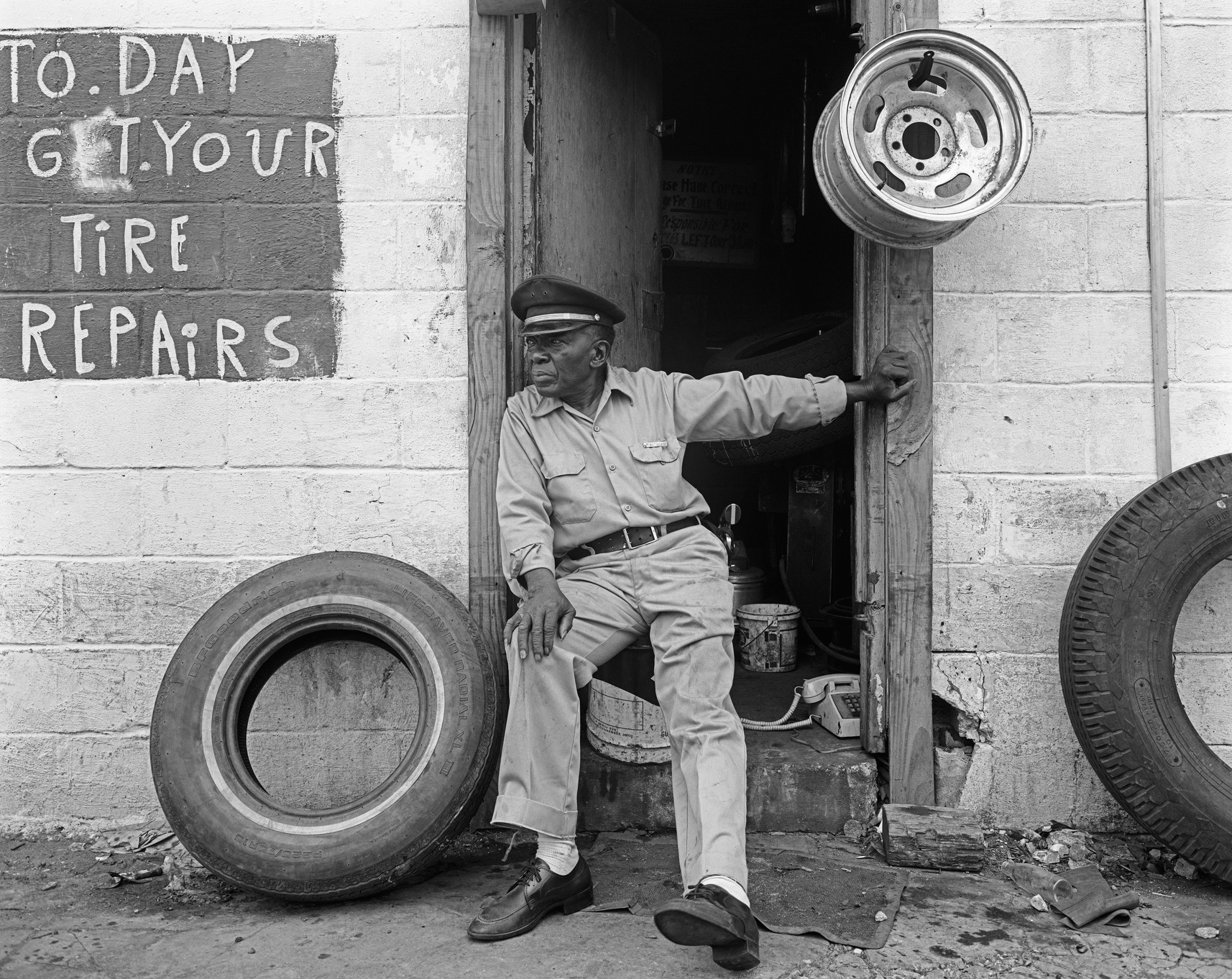 Man sits on a stoop between two tires. painted on the side of the building is a sign that reads, "To. Day Get. Your Tire Repairs"
