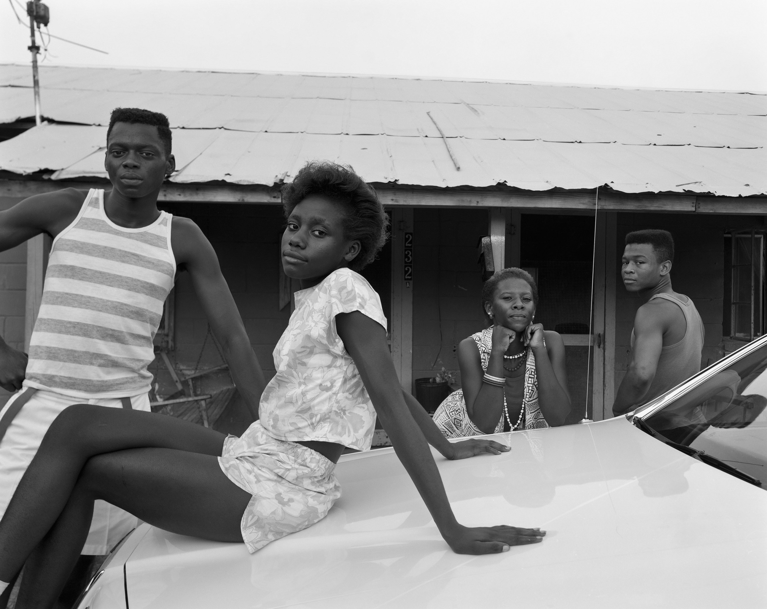 A young woman sits on the roof of a car. Behind her leaning against the same car are also two young men and a woman.