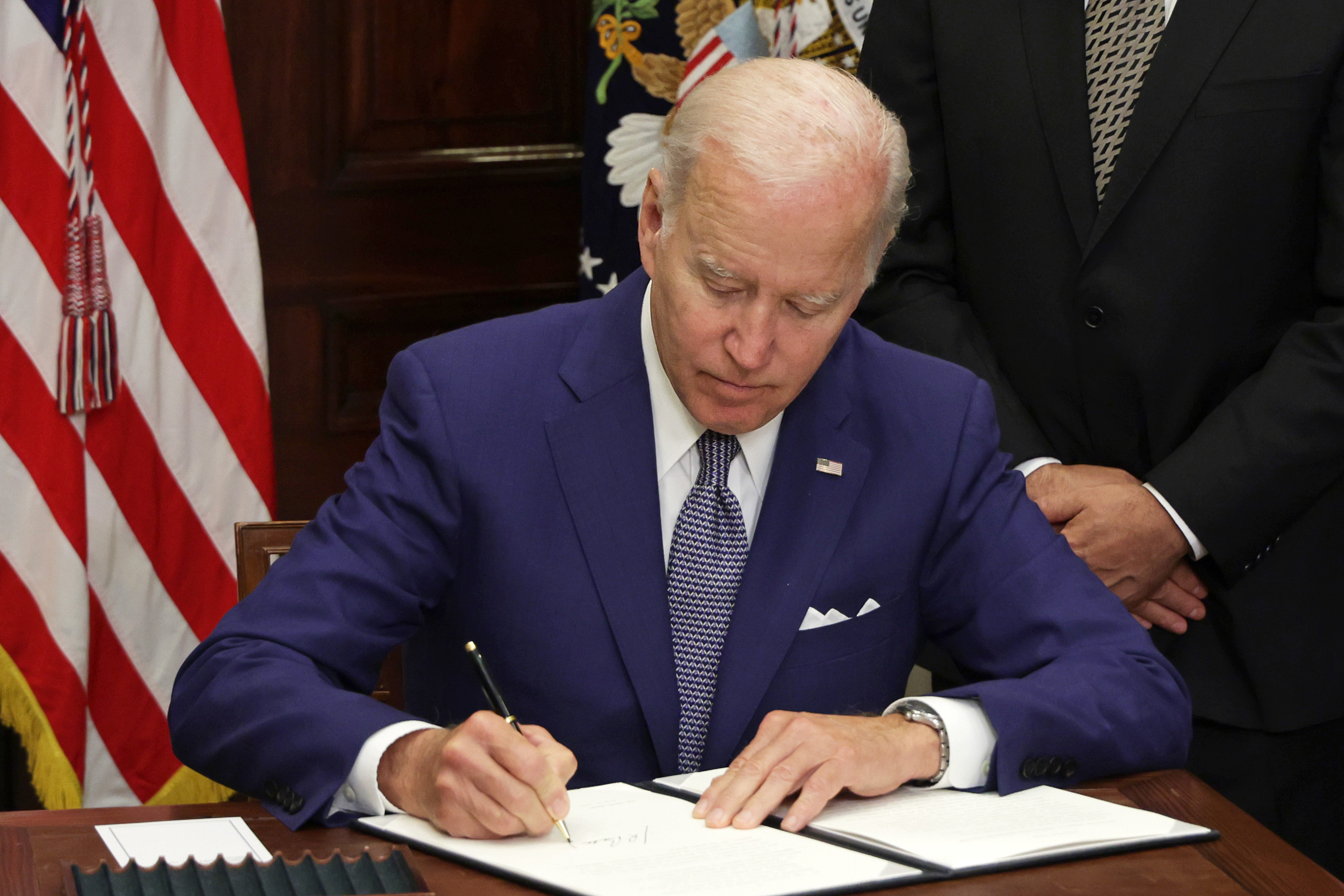 President Joe Biden sit in front of a desk and signs an executive order.