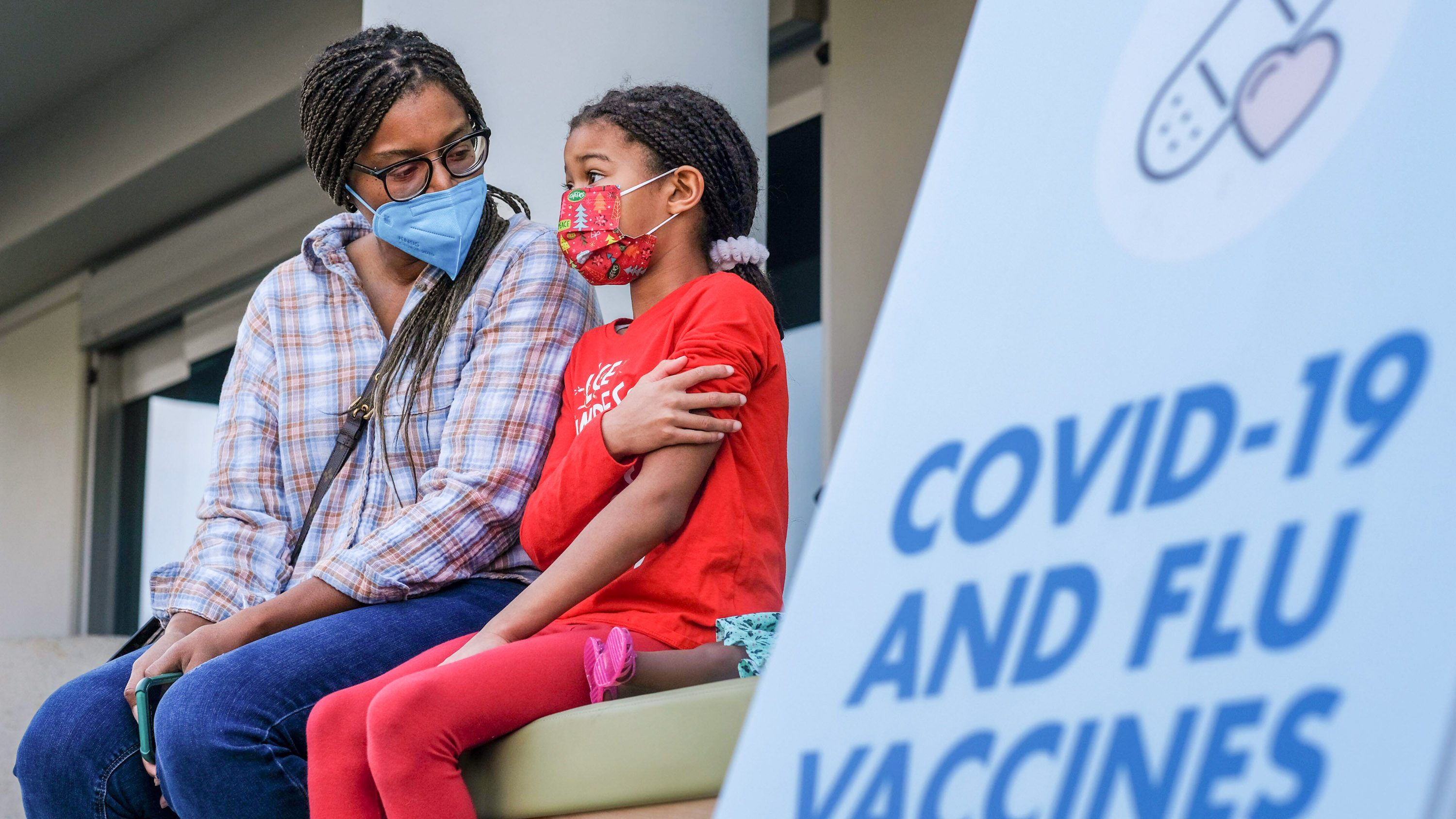 Amelle Samuel, 7, and her mother Kimberli seat in the waiting area after getting a COVID-19 vaccination shot. A sign for COVID-19 and flu vaccines is out of focus in the foreground.