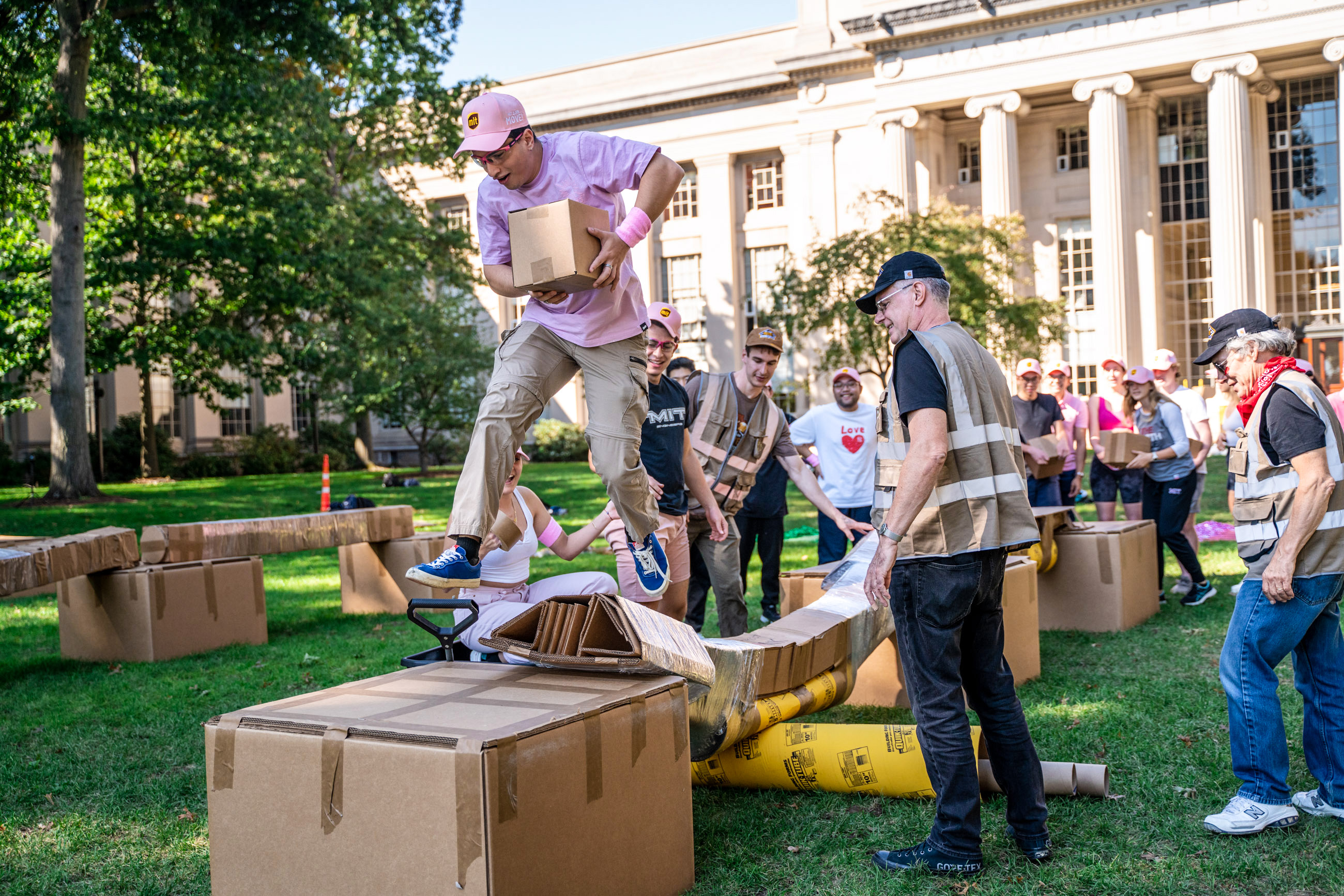 A student is crossing a cardboard brige on the campus lawn while other students watch.