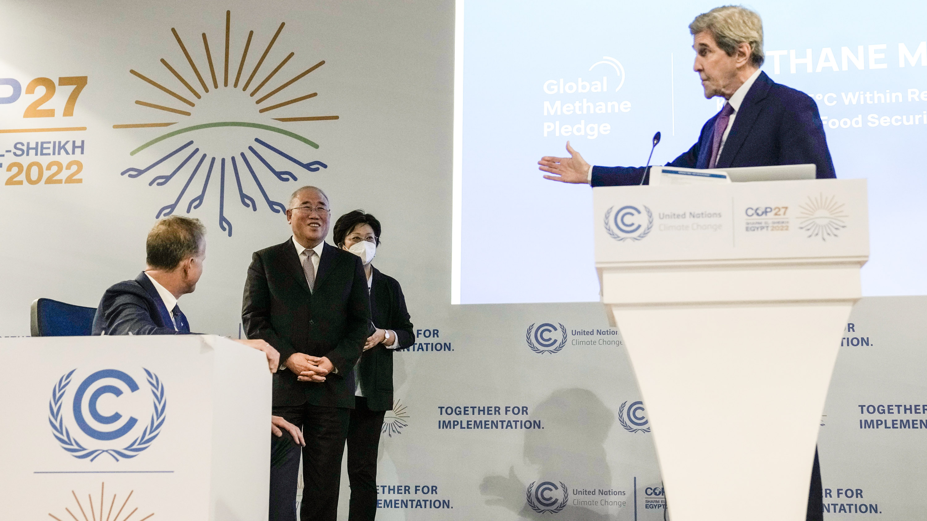 John Kerry at podium of COP27 gestures back toward a smiling Xie Zhenhua.
