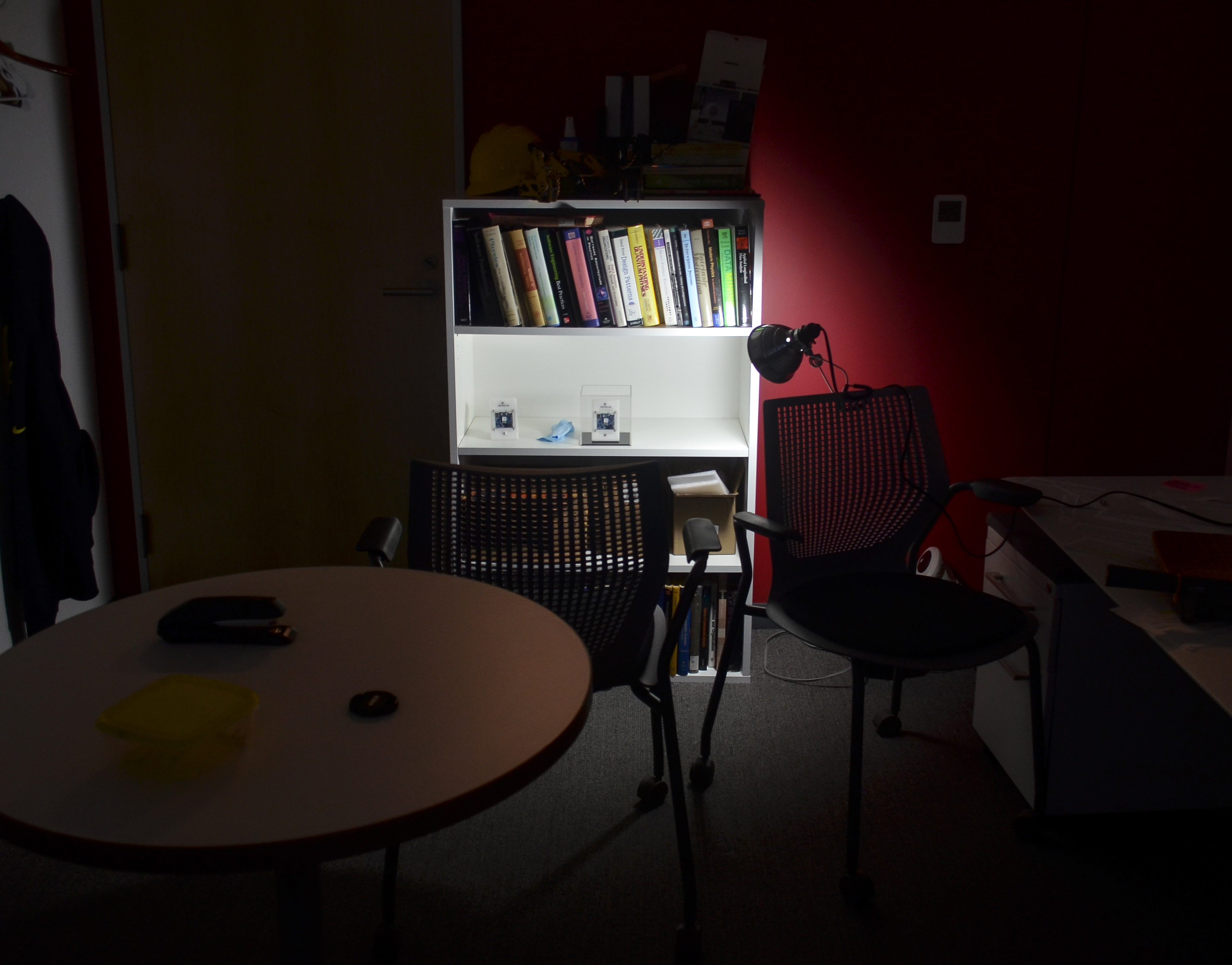 office lit by a single light that is shining on an disconnected Mites panel on a shelf that is housed in a plexiglass cube.