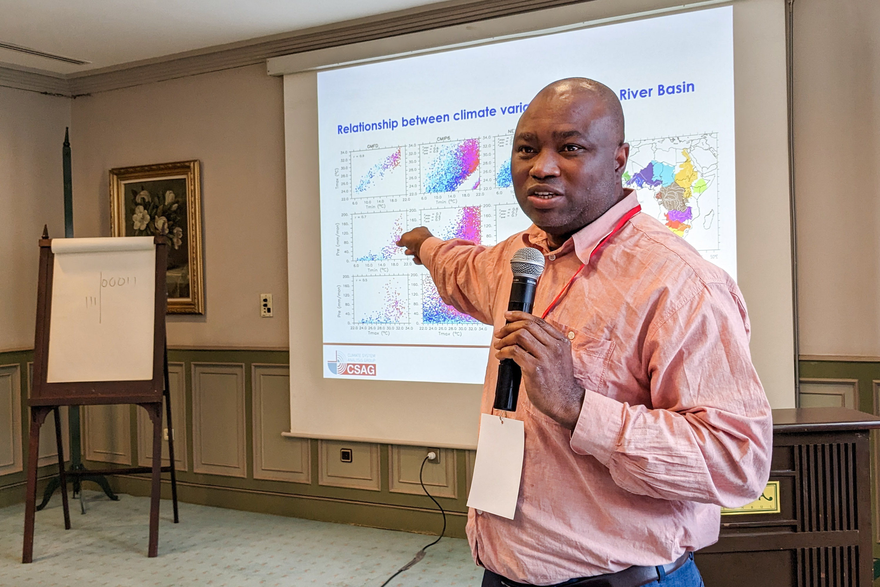 El profesor Abiodun sostiene un micrófono y señala las estadísticas climáticas proyectadas en la pantalla detrás de él.