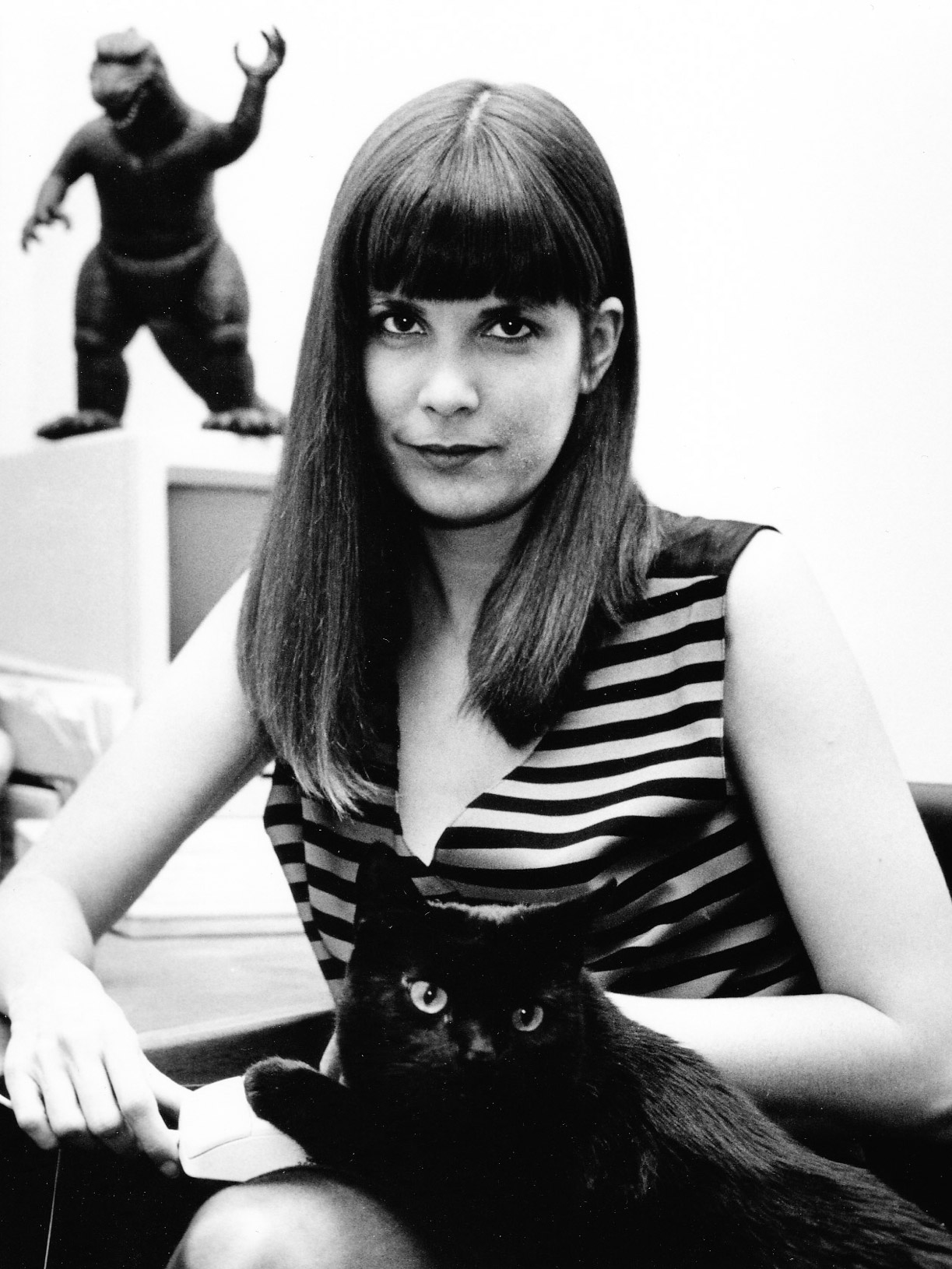 Stacy Horn posing next to a computer with a black cat and a figurine of Godzilla.