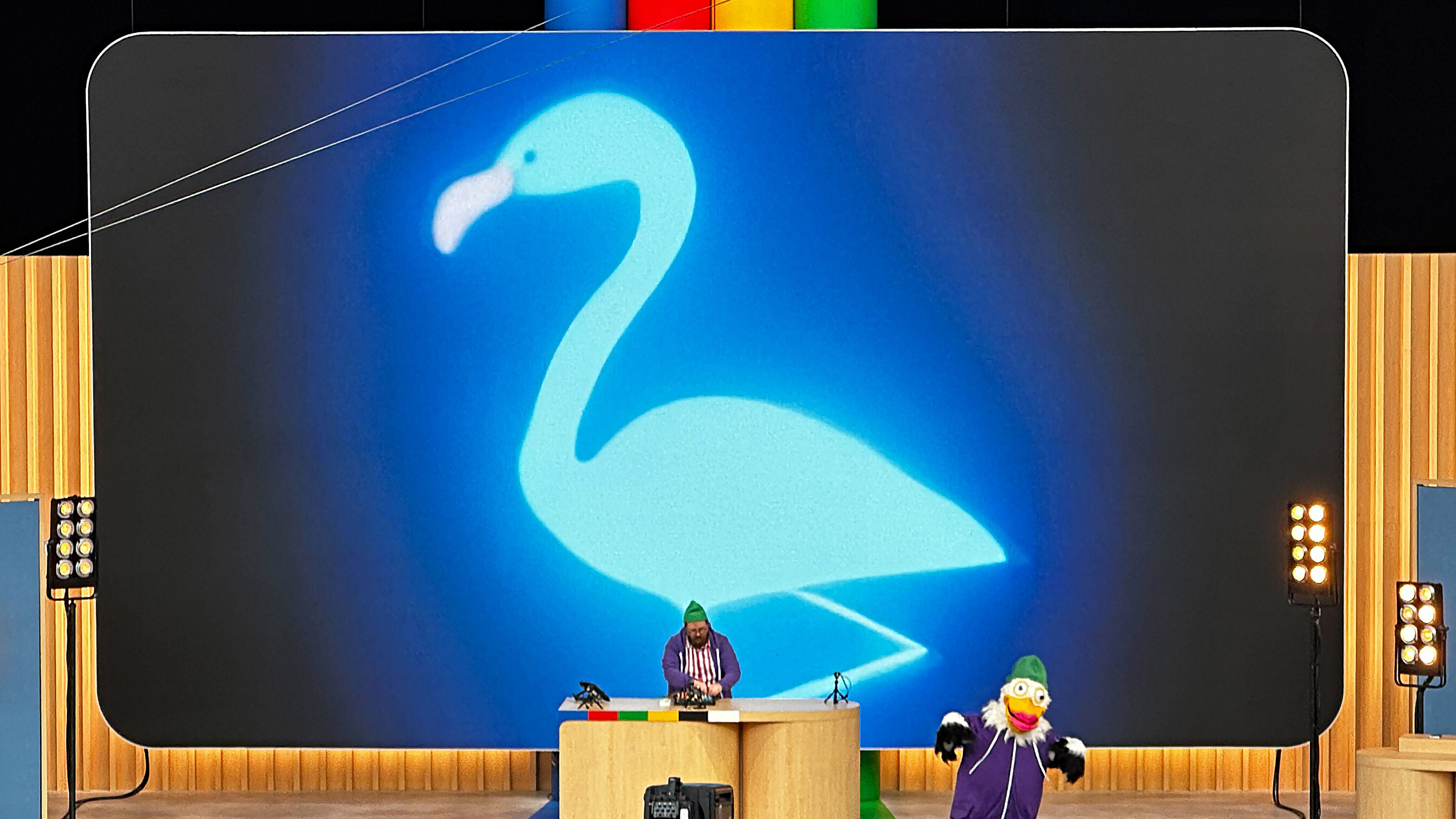 view of stage from audience where Dan Deacon performs as DJ in front of a screen with a giant blue flamingo. A person in a Duck costume is dancing to the right of him.