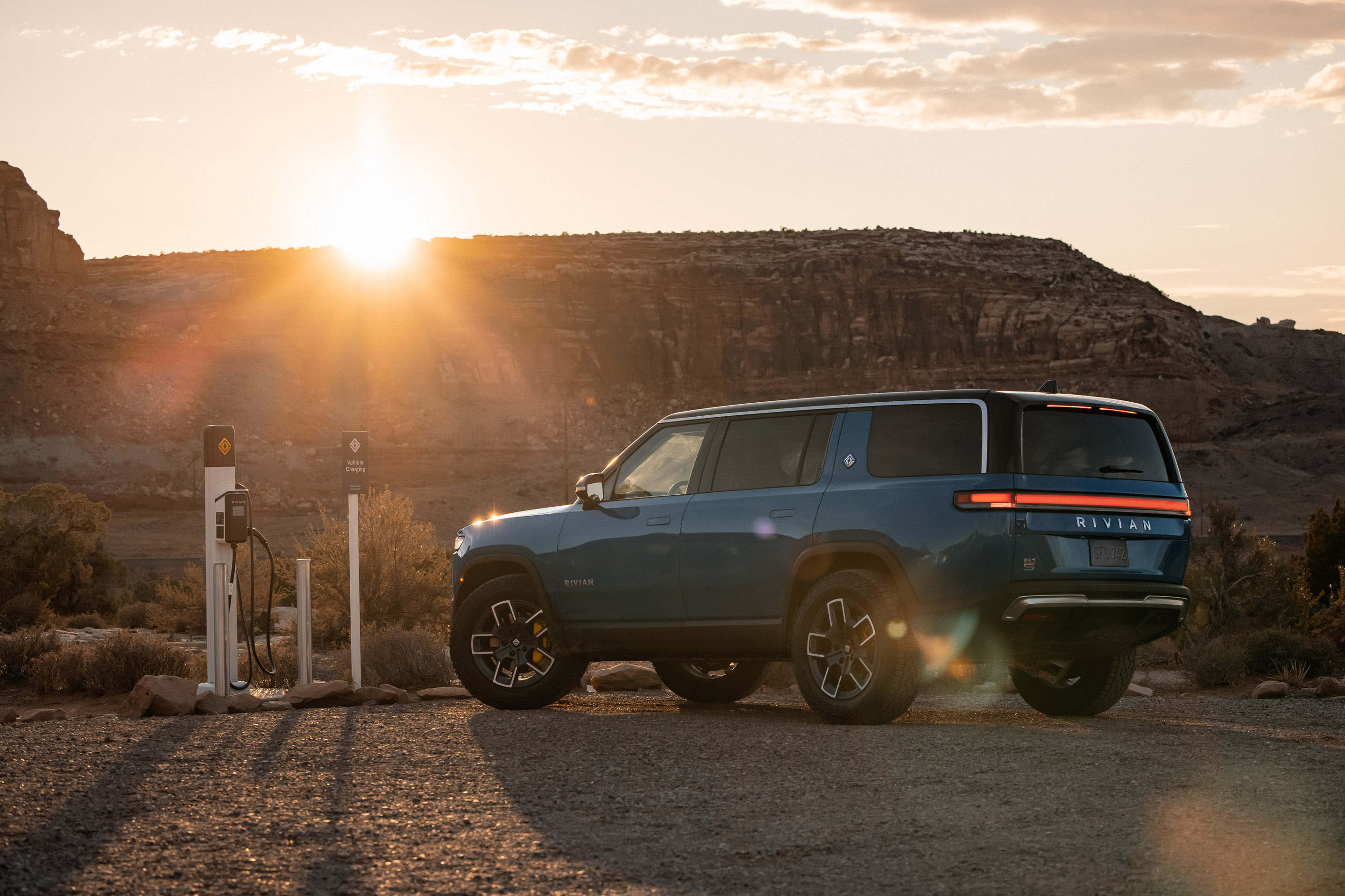 a 2022 Rivian R1S parked next to a charging station in a rugged environment. The sun flares over the edge of a distant mesa.