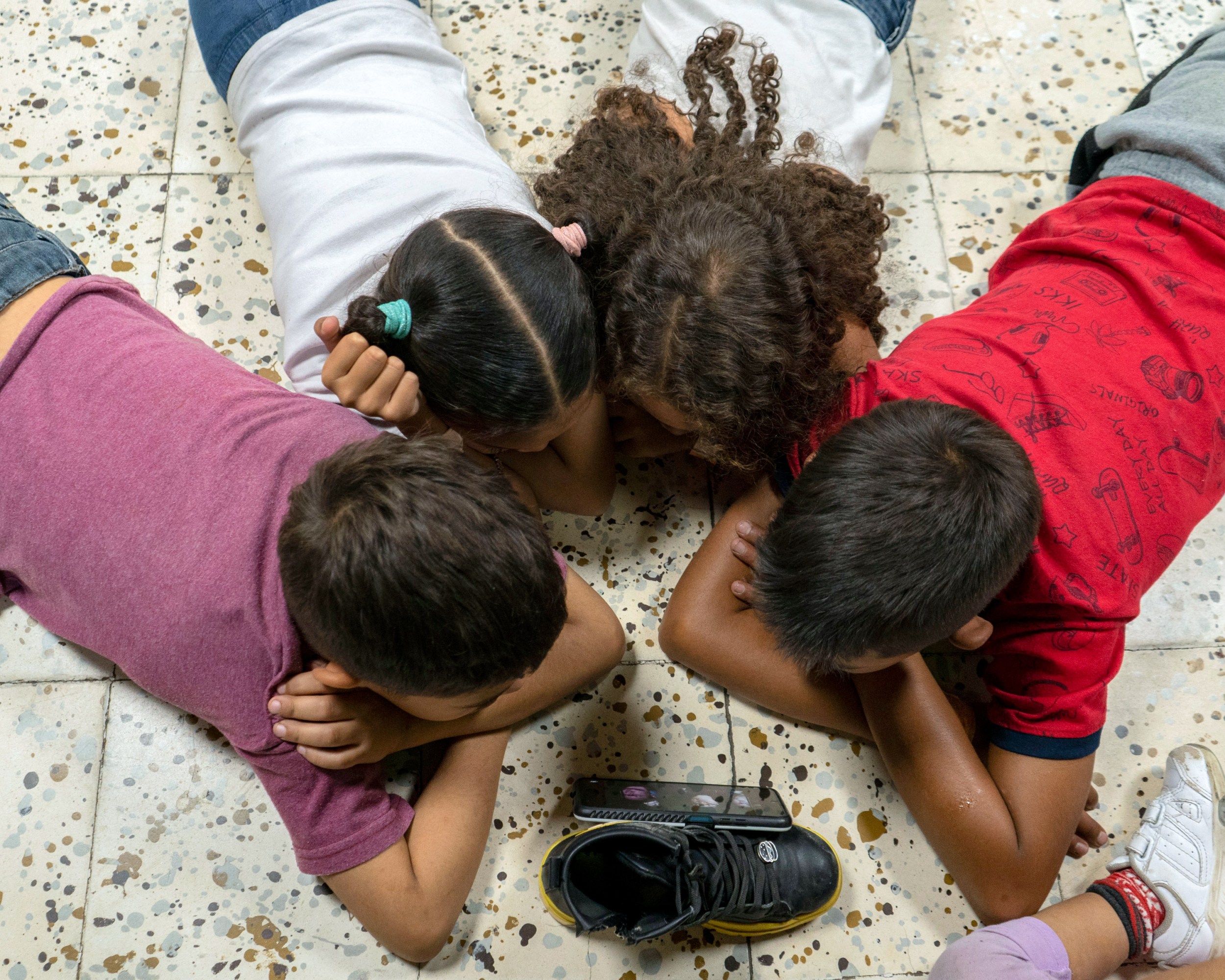 Four children lay on their stomachs on the floor around a cell phone propped up against a shoe.