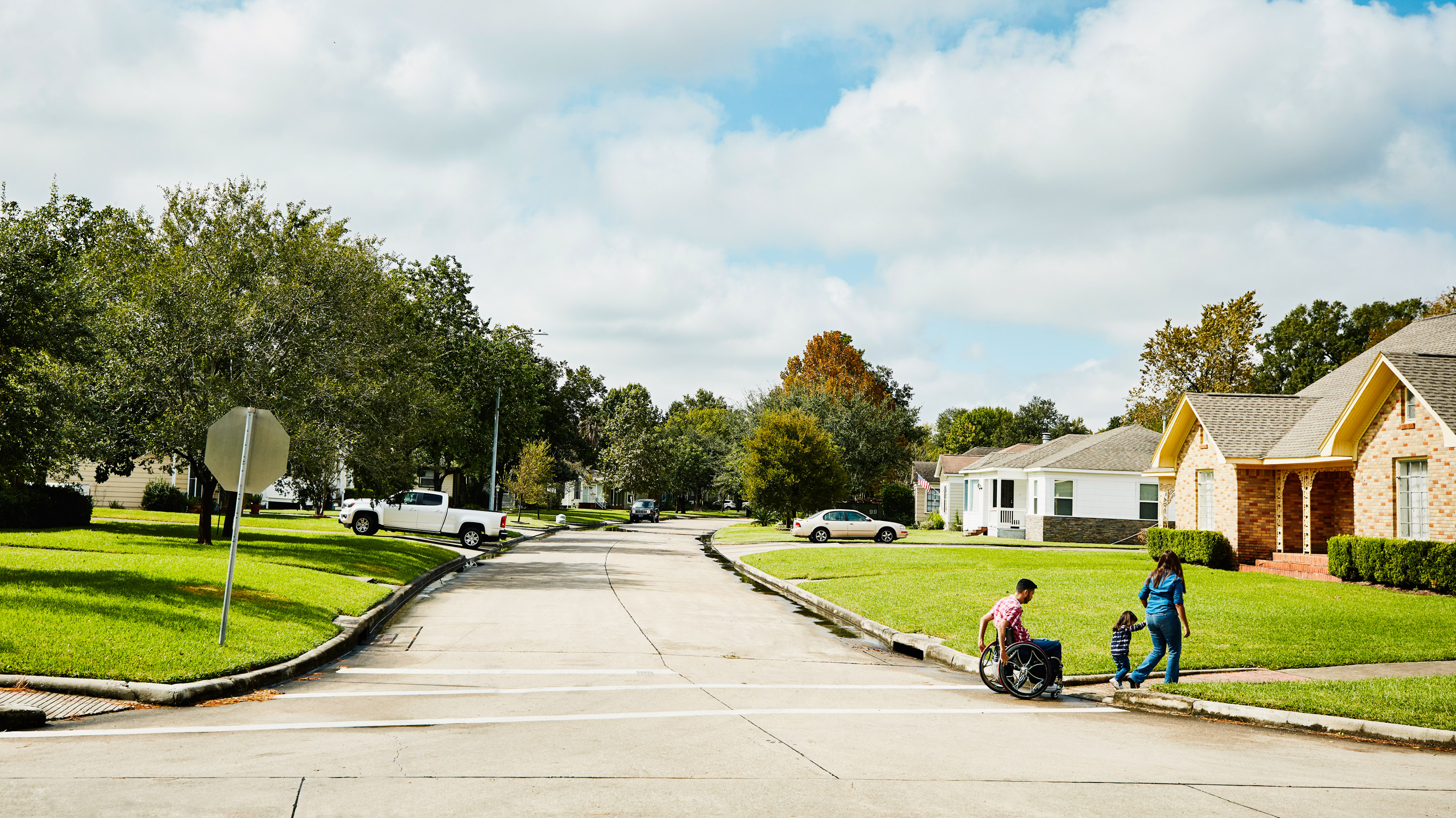 Looking down a neighborhood street where a man in wheelchair has crossed with wife and daughter.