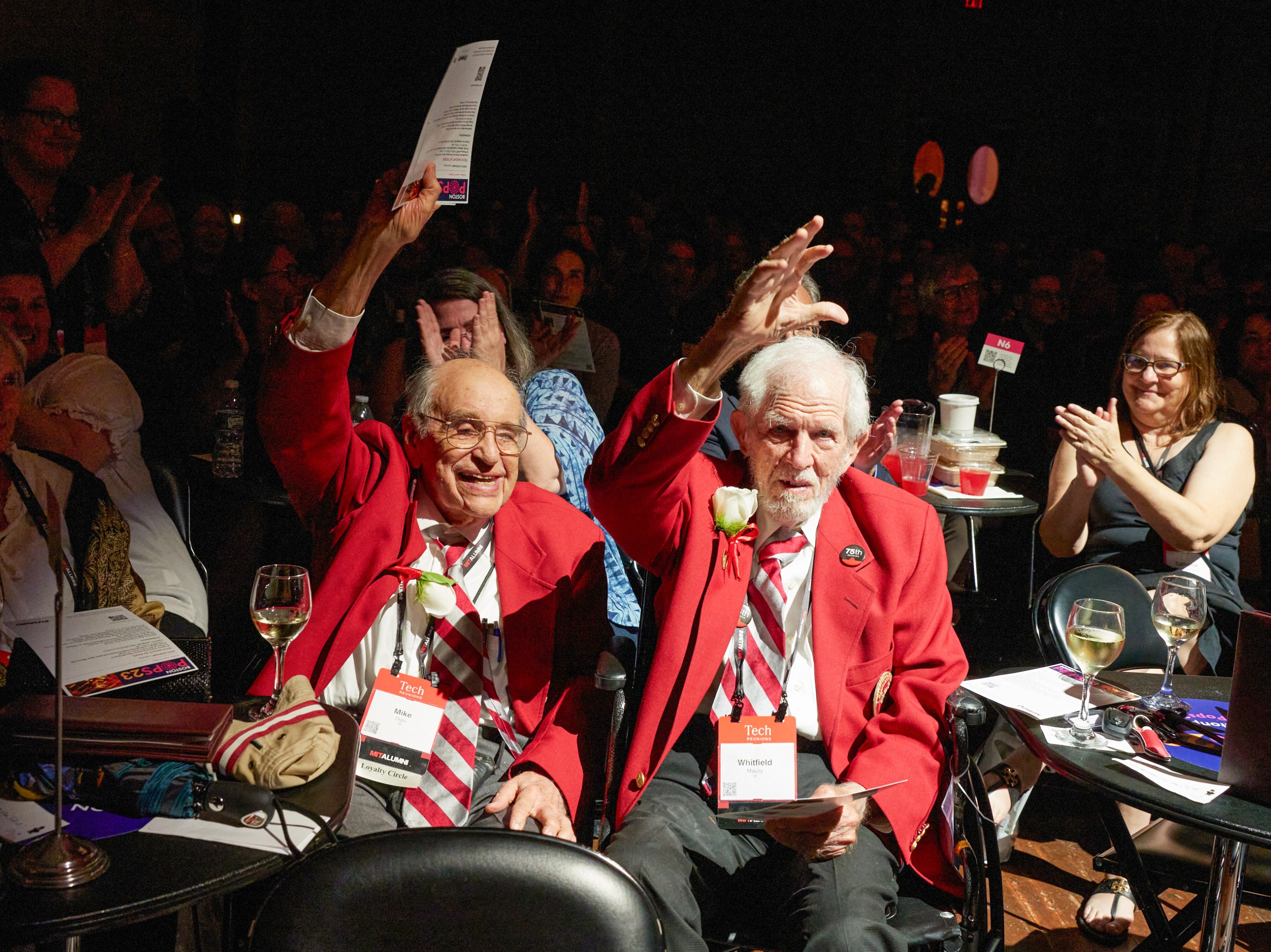 two elderly men in MIT jackets wave at the camera