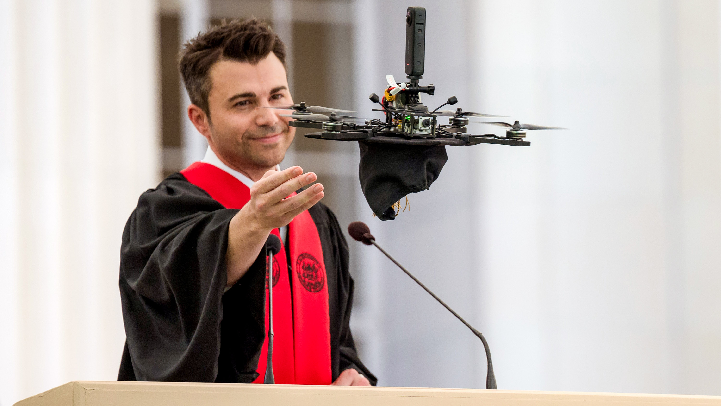Mark Rober at the commencement podium with one hand outstretch toward a drone hovering in the air just in front of him