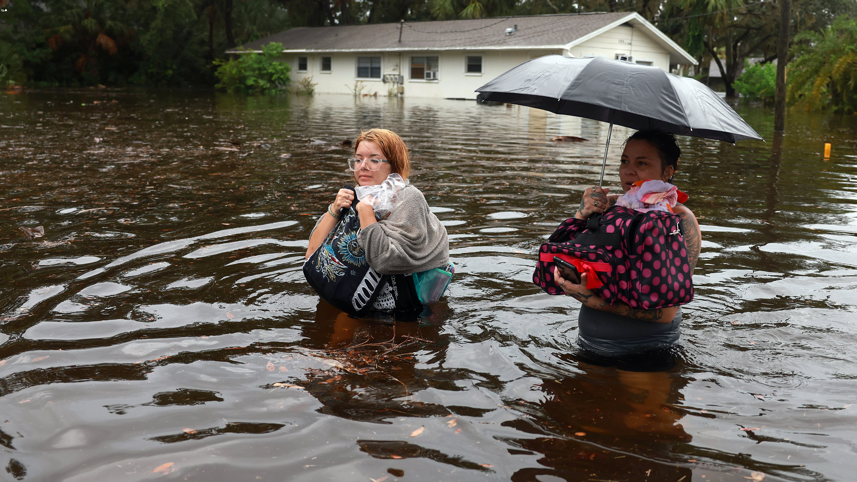Two women wade through waist-high water holding up their belongings.