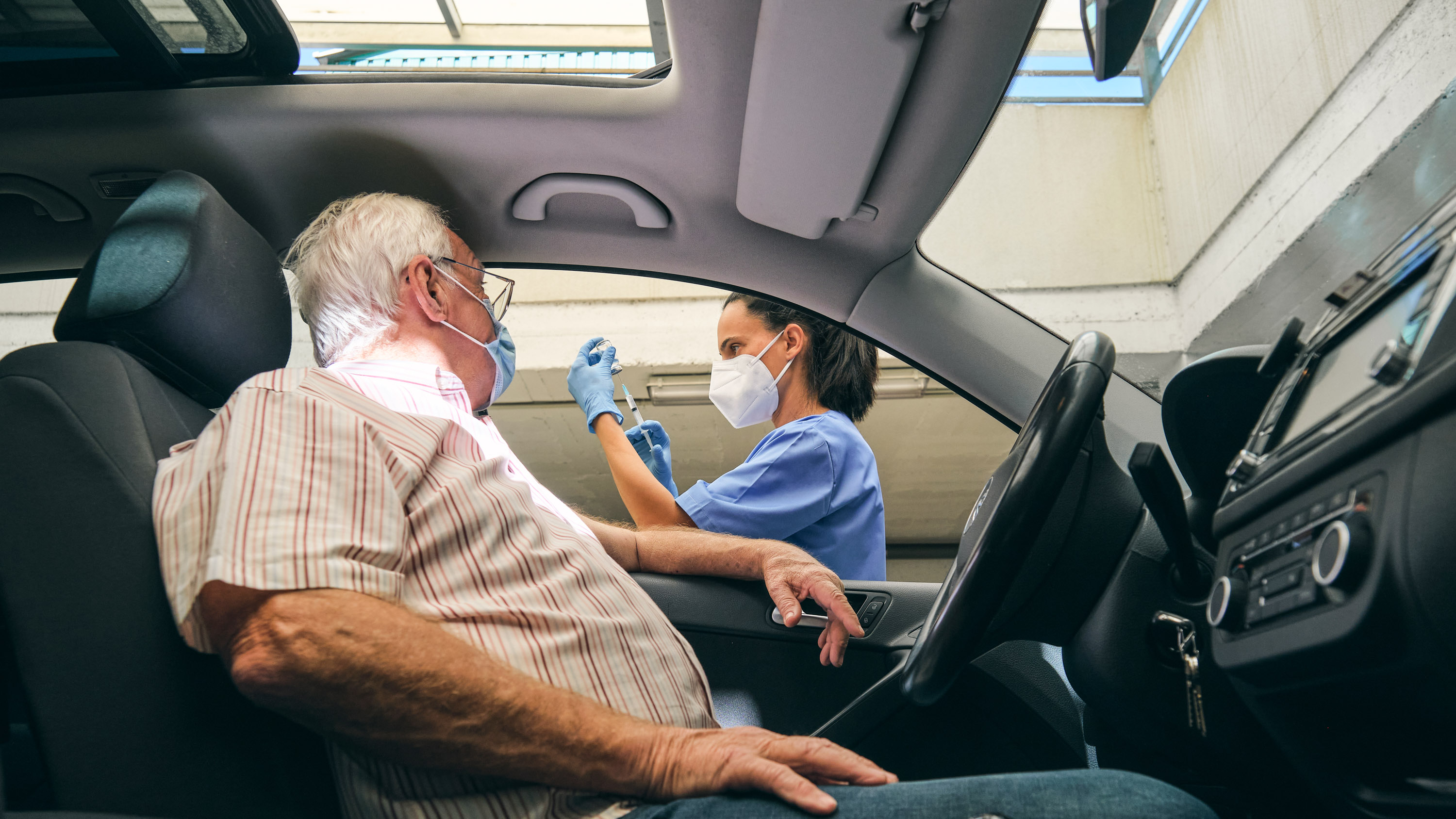 Elderly male patient in automobile watches a medical professional prepare his covid vaccine at a drive-thru clinic
