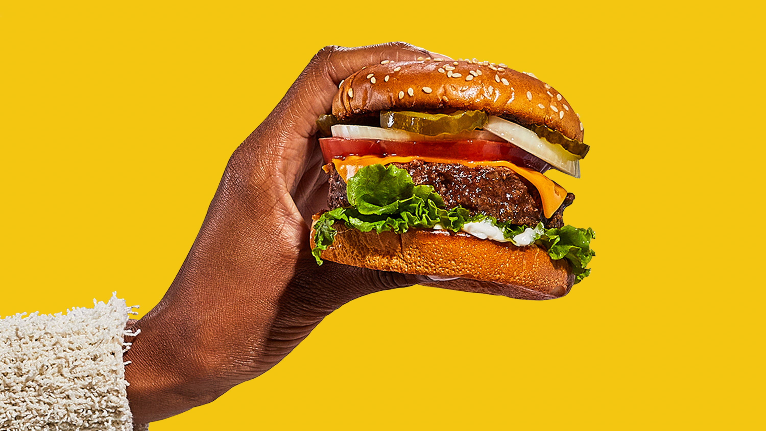 a hand holds a burger in a seeded bun with pickle, lettuce, tomato, onion and cheese.