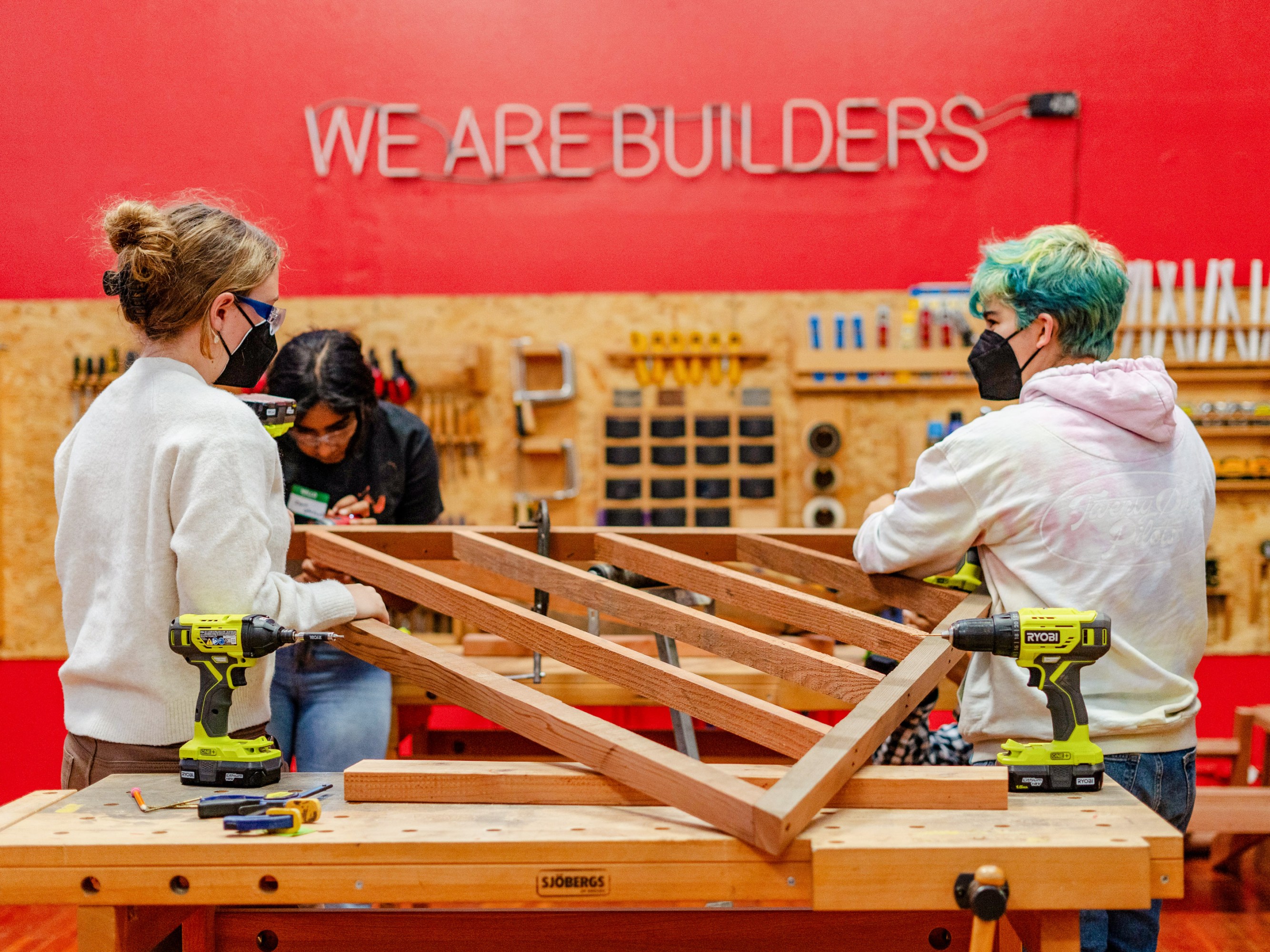 young women with a wooden frame in the Girls Garage. On the bright red painted wall behing them is says "We are Builders" in a neon sign