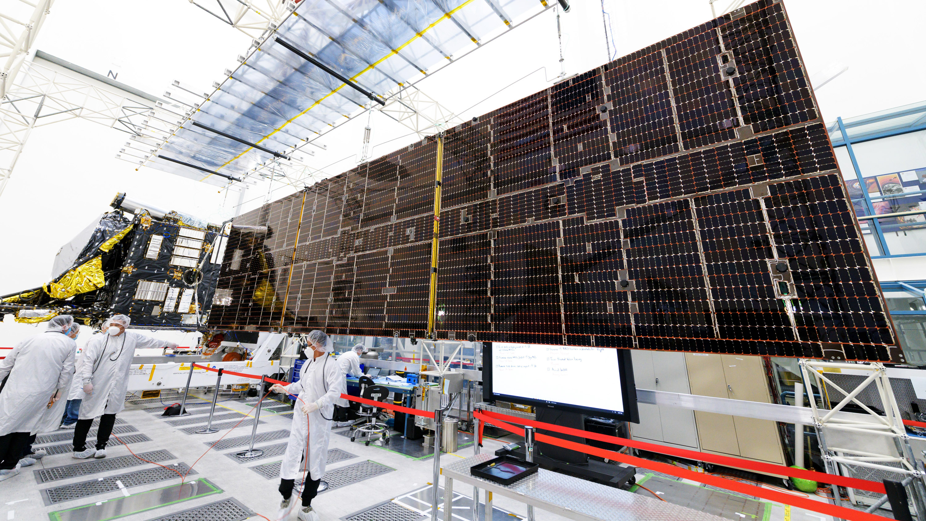 a massive solar array on a frame in the cleanroom with workers in cleansuits standing beneath it