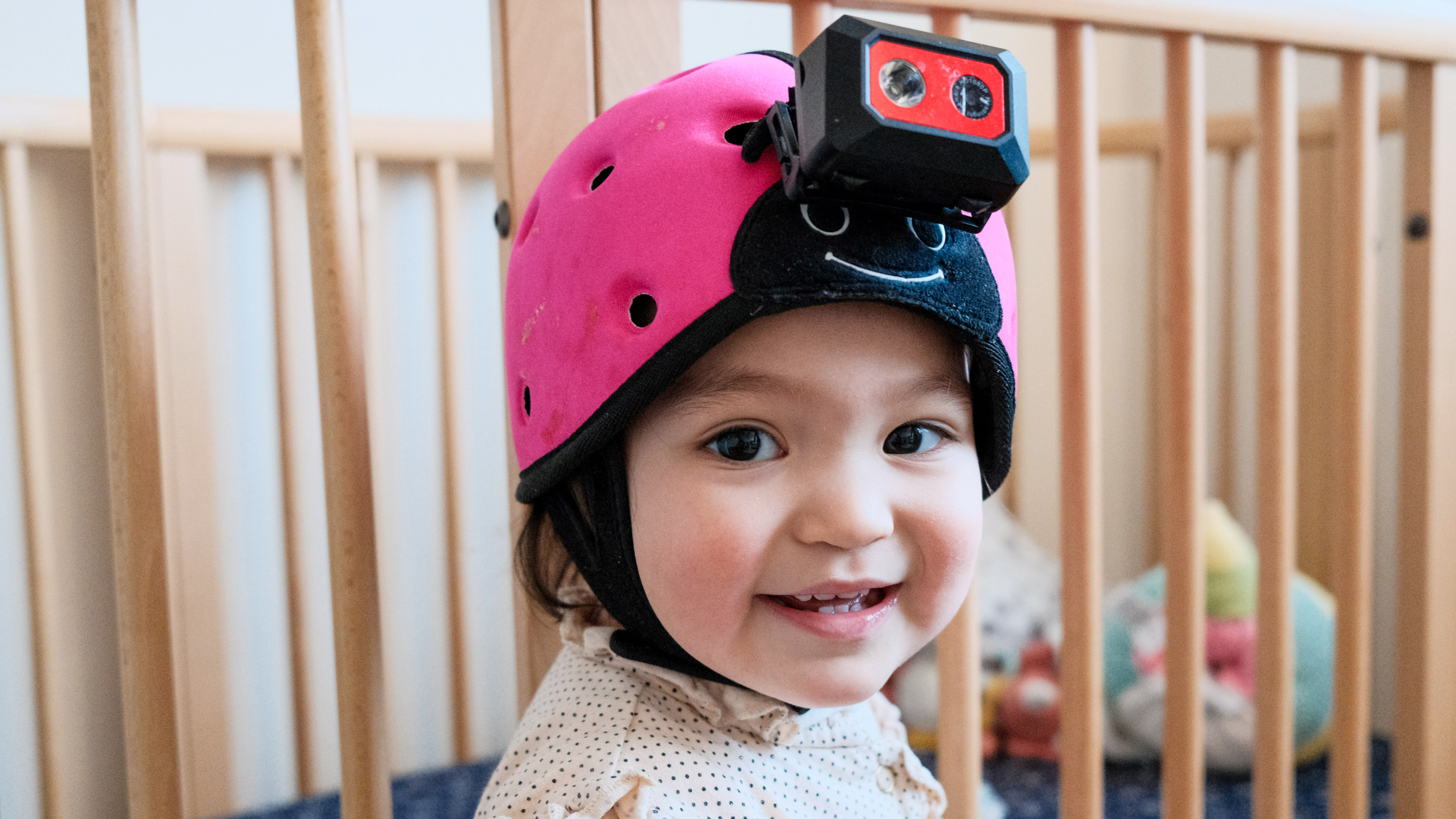Nahaufnahme eines lächelnden Babys, das eine Headset-Kamera trägt, mit den Gitterstäben eines Kinderbetts im Hintergrund