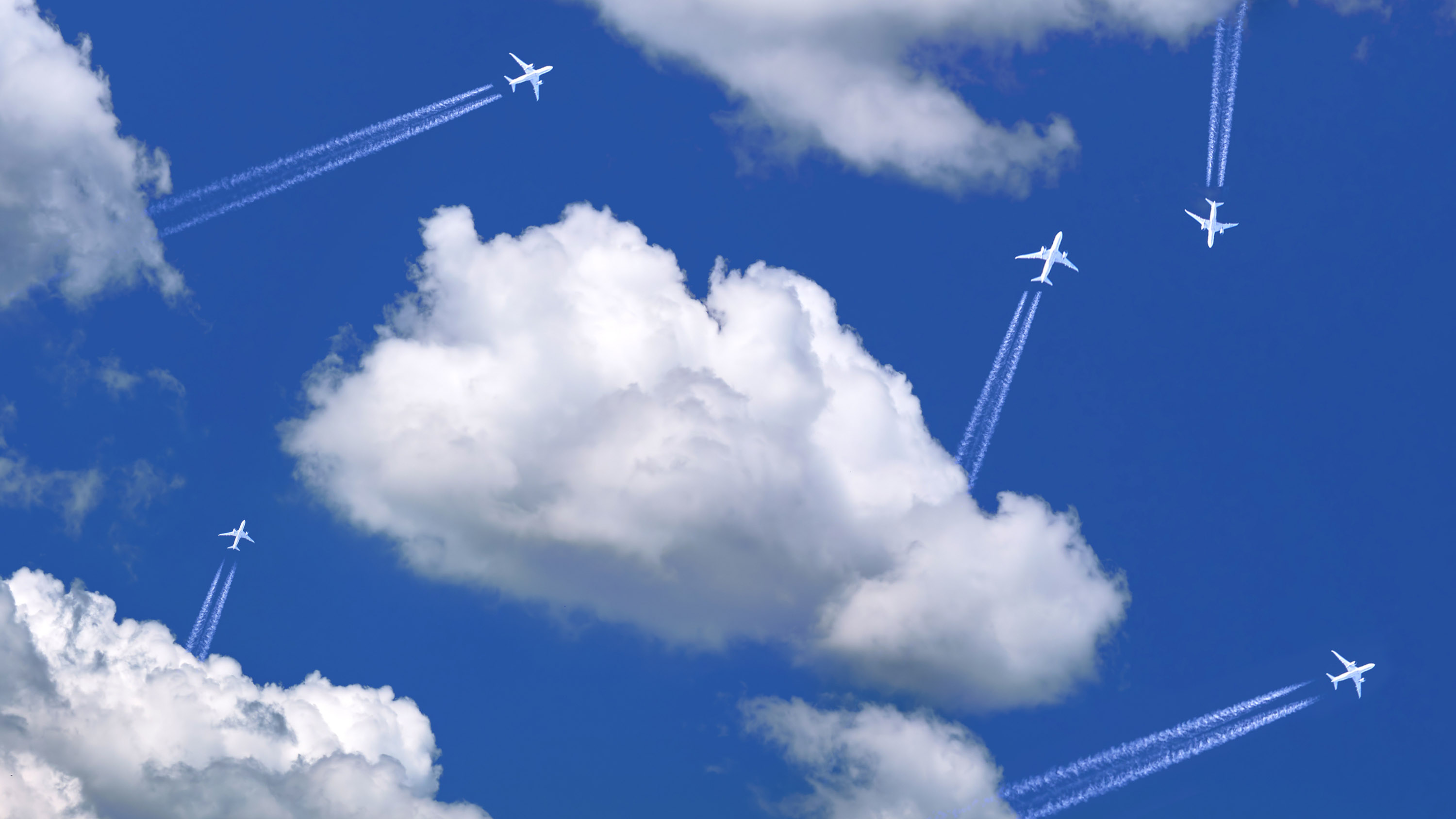 five planes flying out of white puffy clouds at different angles across a blue sky, leaving contrails behind