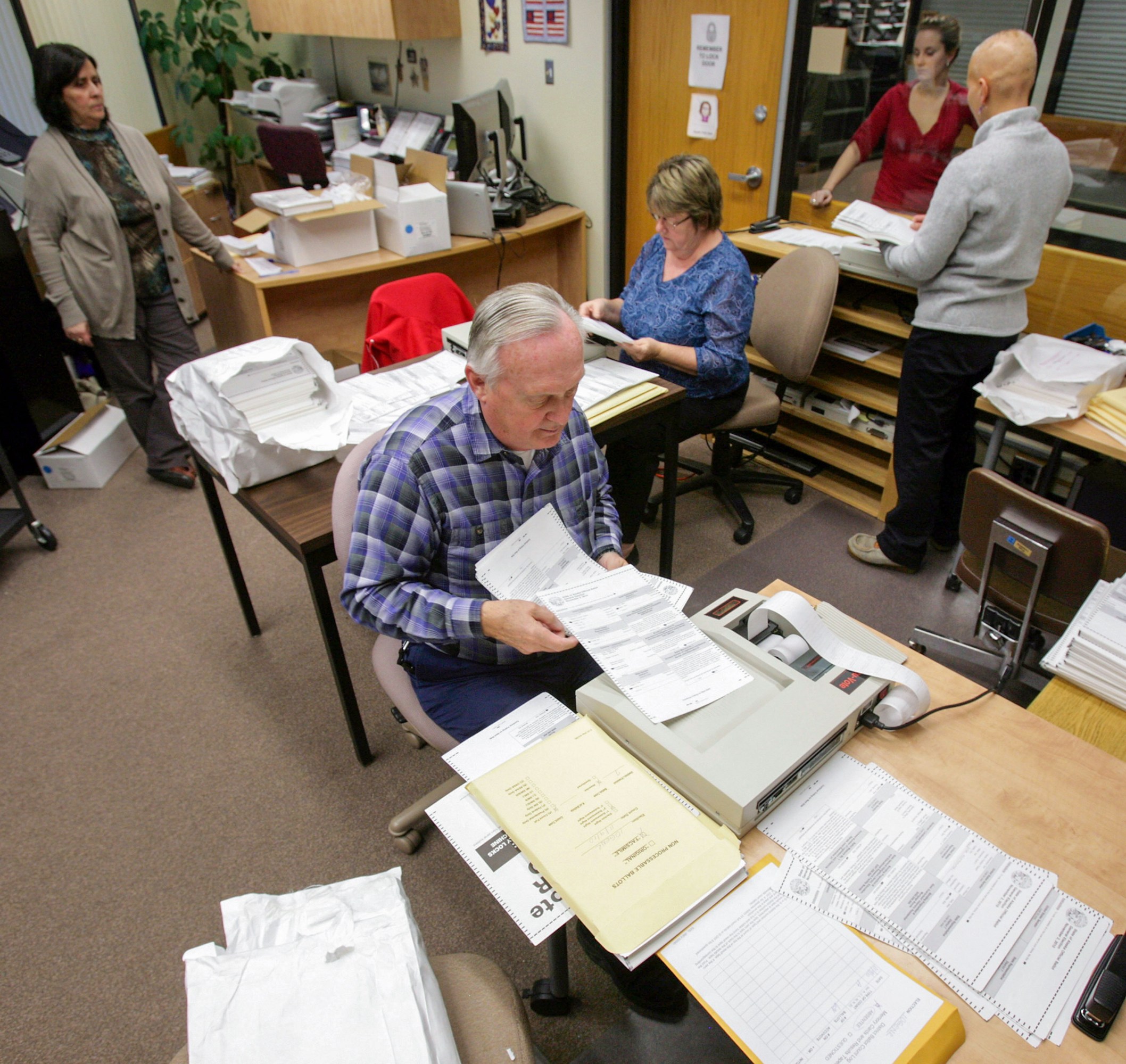 people in an office setting surrounded by stacks of ballots