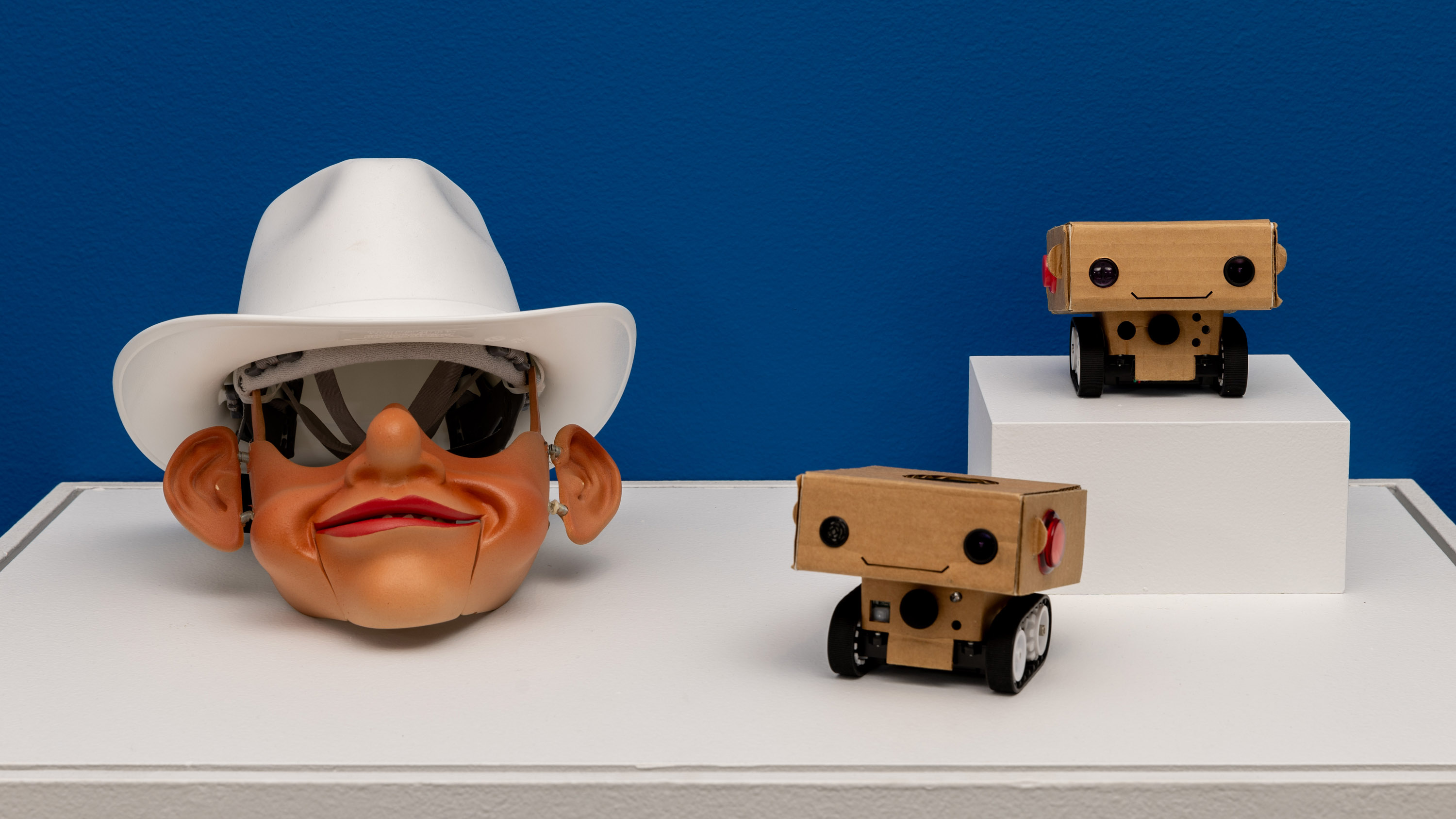 three small sculptures on a white plinth. The first is a puppet head wearing a white cowboy hat and the other two are small smiling cardboard robots on plastic conveyor wheels