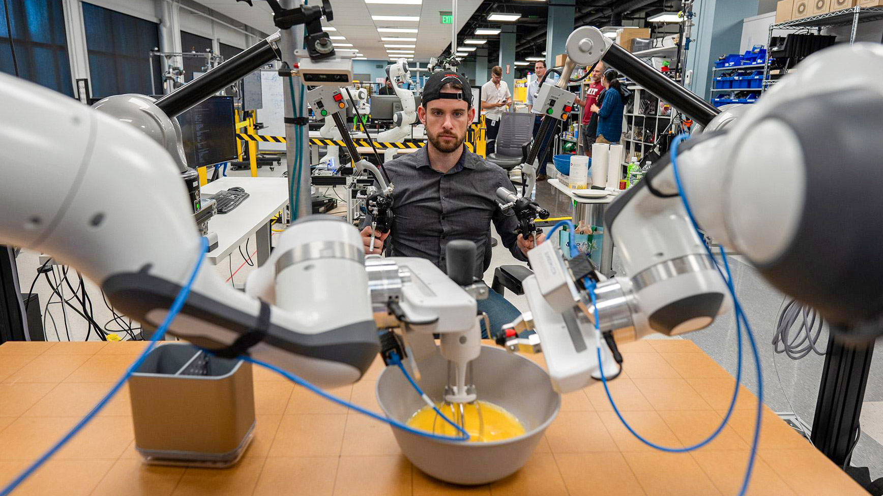 a researcher controls robot arms fitted with whisk attachment beating eggs in a workshop environment
