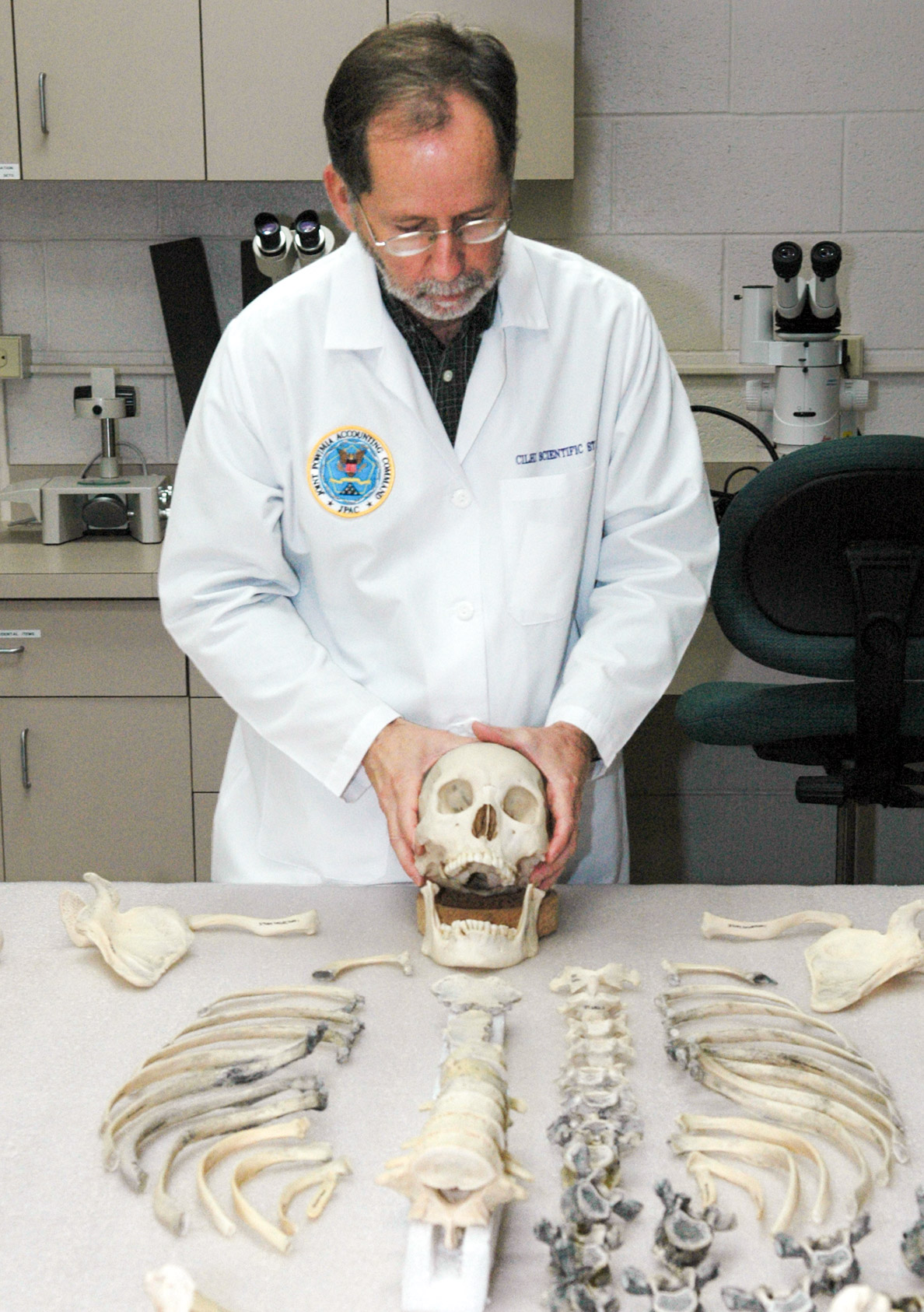 Rober Mann con bata de laboratorio y un esqueleto humano sobre la mesa frente a él.