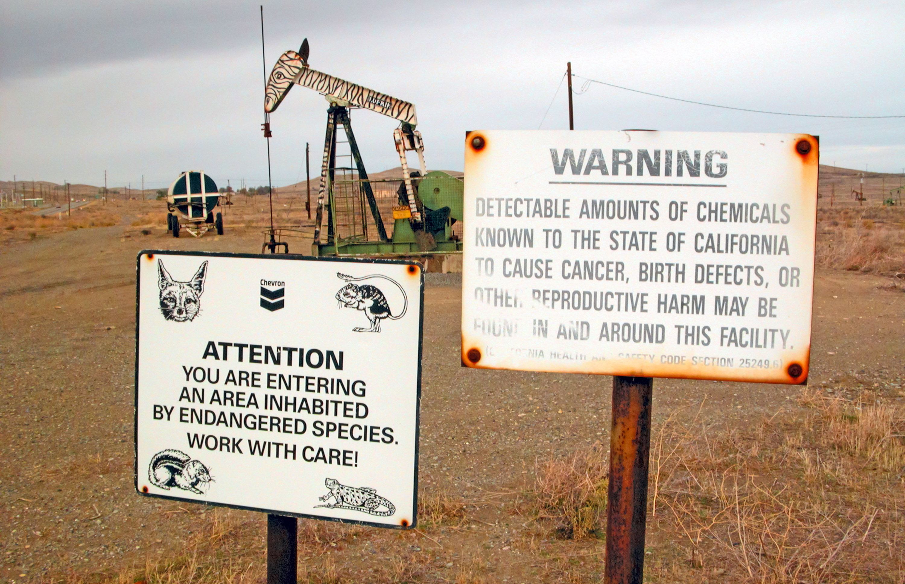 An oil field with a pump jack behind two signs reading "Chevron. Attention you are entering an area inhabited by endangered species. Work with care!" and "Warning. Detectable amounts of chemicals known to the state of California to cause cancer, birth defects, or other reproductive harm may be found in and around this facility."