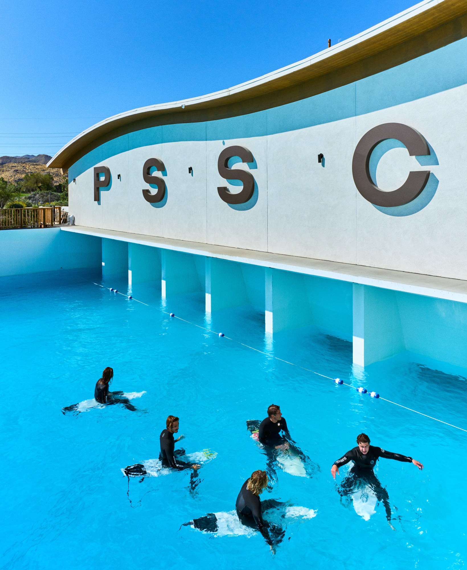 Five surfers sit on their boards in a calm PSSC pool